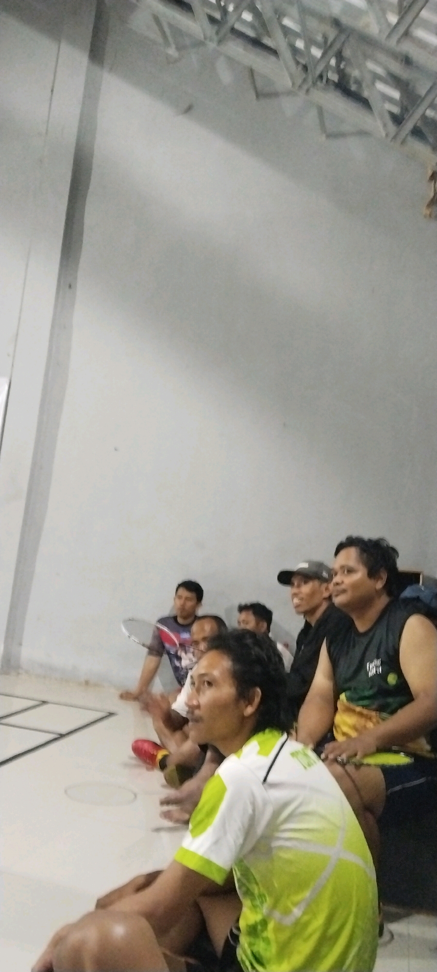 In Kliris, Indonesia, a group of men are gathered indoors, possibly in a sports facility. The lighting suggests an indoor environment, with fluorescent lights visible on the ceiling. The floor appears to be a light-colored surface, possibly linoleum or concrete, marked with dark lines forming rectangular sections.
Several men are seated, with at least three in the foreground. The man closest to the camera is wearing a white and lime green t-shirt with a geometric pattern. He has dark, wavy hair and is looking towards his left. To his right, another man is visible, wearing a black sleeveless shirt and a yellow and green patterned sarong or sash. He is looking to his left and has a serious expression. Further to the left, a man in a black t-shirt and a grey baseball cap is smiling and looking to his right. Behind him, another man in a black shirt and a cap is partially obscured, looking to his left. To the far left, a man in a patterned t-shirt is seated and holding a badminton racket. Other individuals are also present in the background, some of whom are looking towards the front or to the side. The overall atmosphere appears to be relaxed, with people engaged in conversation or watching something.