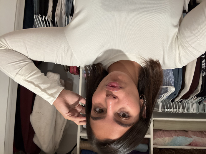 An adult female is depicted in a close-up selfie, posing indoors within what appears to be a closet or dressing area. She has dark, shoulder-length hair, a tanned complexion, and is wearing a light-colored, long-sleeved ribbed top. Her lips are puckered, and her gaze is directed at the camera. Two silver hoop earrings are visible on her left ear. Her right hand is positioned near the right side of her head. The background consists of an organized closet interior featuring multiple white shelves and hanging rods. The left side displays folded textiles in various colors, including pink and blue, on upper shelves, and numerous garments on white hangers below. The right side reveals a white, fluffy coat hanging, a dark red garment, and small pink and brown bags resting on a lower shelf. A white door frame is partially visible on the far right. The specific location in the city of Princeton, United States, cannot be determined from the provided image.
