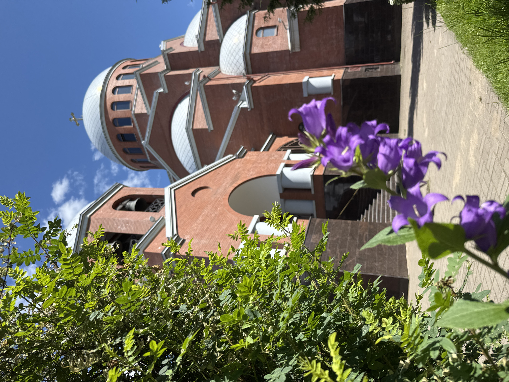 This image depicts the Holy Trinity Cathedral in St. Petersburg, Russia, on a clear, sunny day with a bright blue sky and scattered white clouds. In the foreground, out of focus, are vibrant purple bell-shaped flowers, with green leaves and branches of a bush obscuring the left side of the frame.
The cathedral itself is constructed of red brick and features several domed roofs, one large central dome topped with a golden cross, and a prominent bell tower. The architecture is ornate, with arched windows and white columns adorning the entrance. The grounds surrounding the cathedral appear to be paved with light-colored brickwork, and a strip of green grass runs along the right edge of the image. The scene is serene and bathed in natural light.