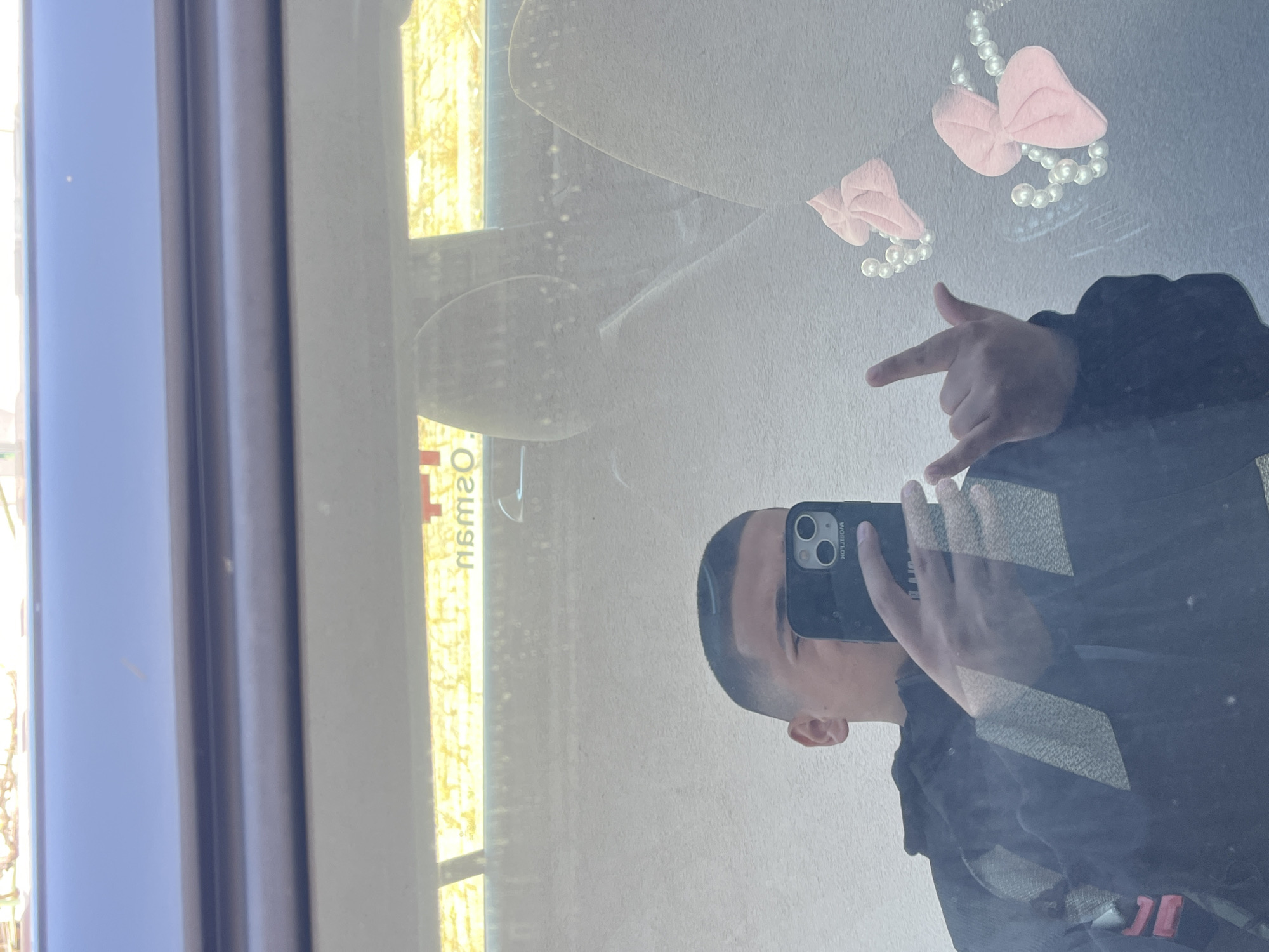 A person takes a selfie in front of a window displaying an advertisement in a shop window. The advertisement features pink hair bows connected by strands of pearls. The location is Lozarevo, Bulgaria, based on the sign's reflection in the window. The person is wearing a black jacket.