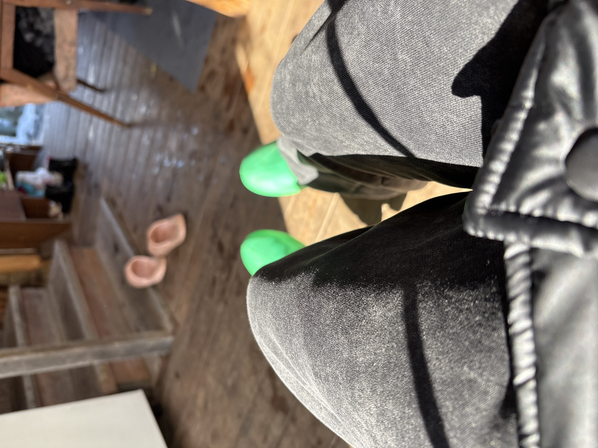 The image shows a downward-angled view of a person's legs, clad in gray jeans, with bright green clogs on their feet. Part of a black jacket is visible on the right side of the frame. The person appears to be standing on a wooden surface, possibly a porch or deck. In the background, there are wooden stairs leading down, with a pair of light pink clogs resting on one of the steps. The image's location is implied to be in Zheleznodorozhnyi, Russia, based on the prompt's instructions, but this is not verifiable from the image itself.