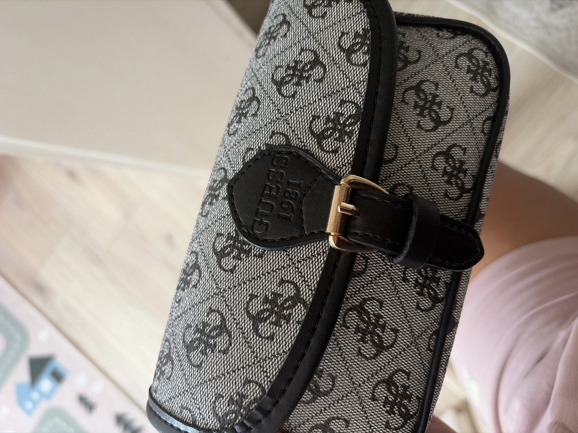 The image shows a close-up of a woman's grey and black patterned Guess handbag. The bag features a prominent "GUESS 1981" logo in black on a leather patch, secured by a gold buckle and strap. The handbag's fabric has a repeating pattern of interlocking "G" logos. The background is blurred, showing a light wooden floor and a pink mat with a cartoon house graphic. The image was taken indoors, likely in a residential setting, with no discernible connection to Duisburg, Germany.