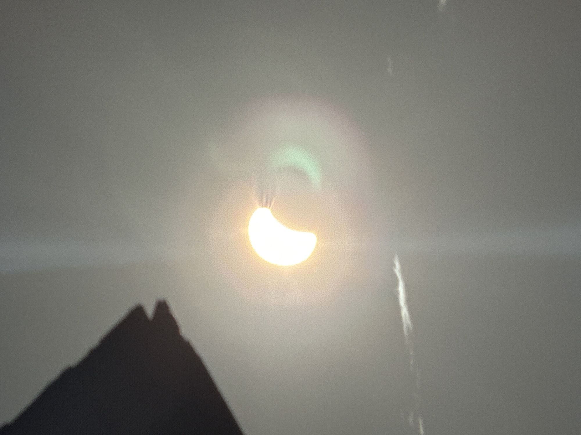 The image depicts a solar eclipse. A dark, triangular shadow is visible in the upper left corner. The sun, partially obscured by the moon, is a bright crescent shape, casting a luminous glow. A faint rainbow-like halo surrounds the sun, with hints of green and pink. The sky appears hazy, with thin, wispy clouds. There are no visible people or specific landmarks to indicate a location in Waterford, Ireland.