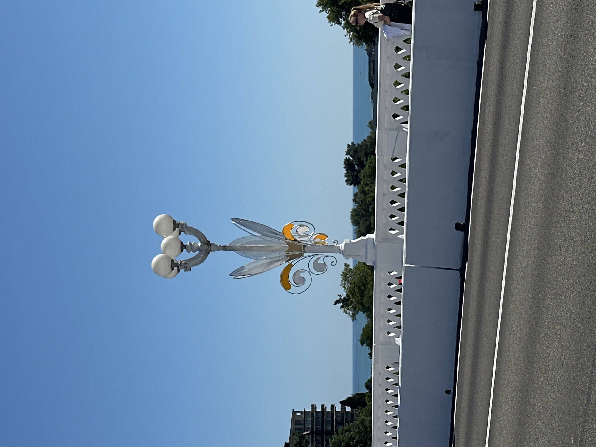 The image captures a view from a bridge, likely in Sochi, Russia, on a bright, sunny day. The primary subject is a decorative lamppost with a unique, artistic design featuring translucent, petal-like elements with yellow accents, and three globe-shaped lights. The lamppost is mounted on a white, ornate balustrade that runs along the side of the bridge.
Beyond the balustrade, a body of water, likely the sea, is visible under a clear blue sky. Lush green trees are interspersed with the scenery, and a modern building with many windows is visible in the distance.
In the upper right corner of the image, a person is leaning over the balustrade, looking out at the view. Their posture suggests they are enjoying the scenery. The overall atmosphere is peaceful and serene, indicative of a pleasant day for sightseeing. The image appears to be taken from a slightly elevated perspective, looking across the bridge and out towards the sea. The asphalt road of the bridge is visible on the right side of the frame, marked with white lines.