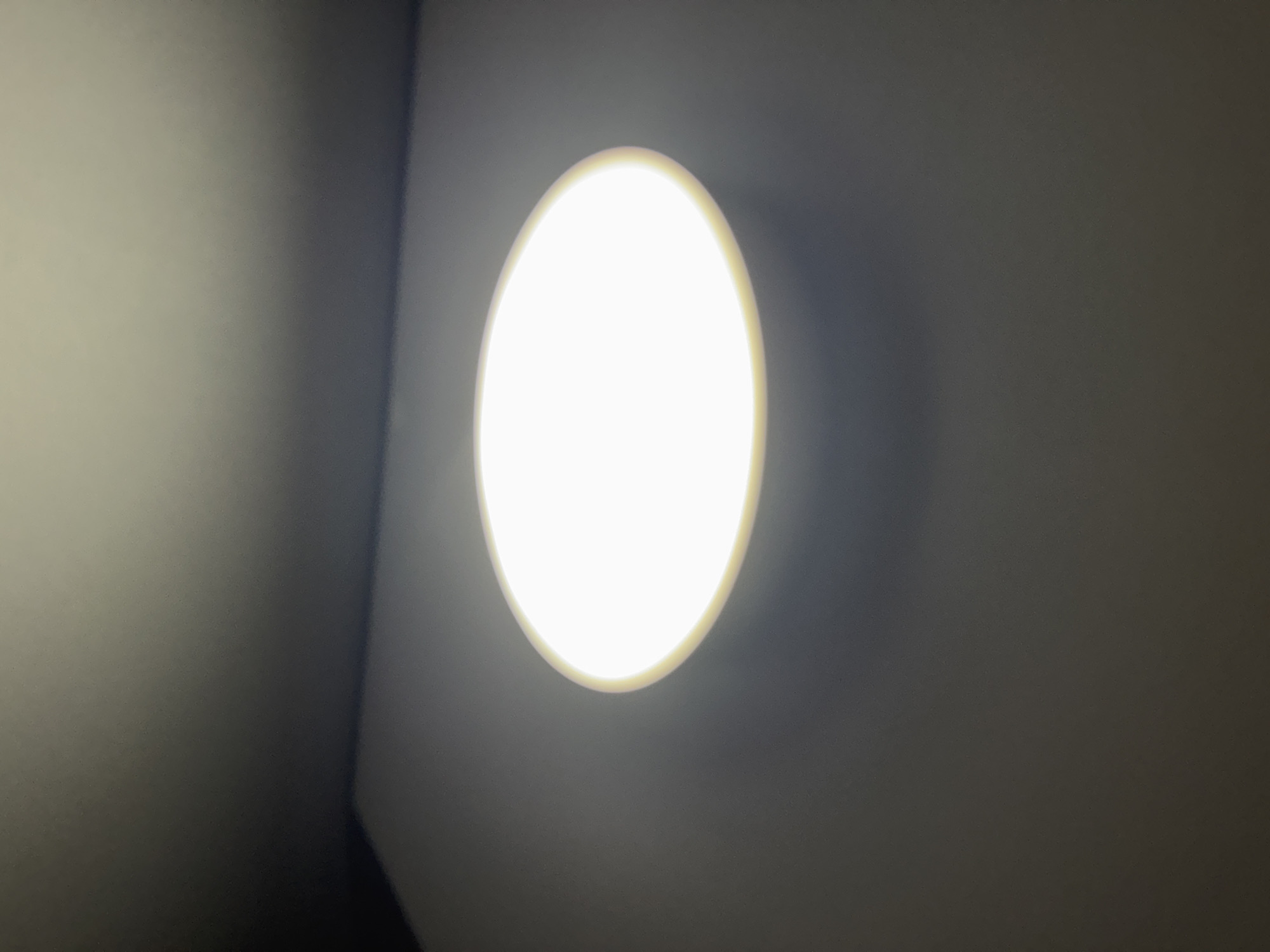 The image shows a light fixture illuminating a dimly lit room. The light fixture is oval-shaped and mounted on the wall. The wall is painted a dark neutral color. The corner of the room is visible on the left side of the frame. The location is unknown.