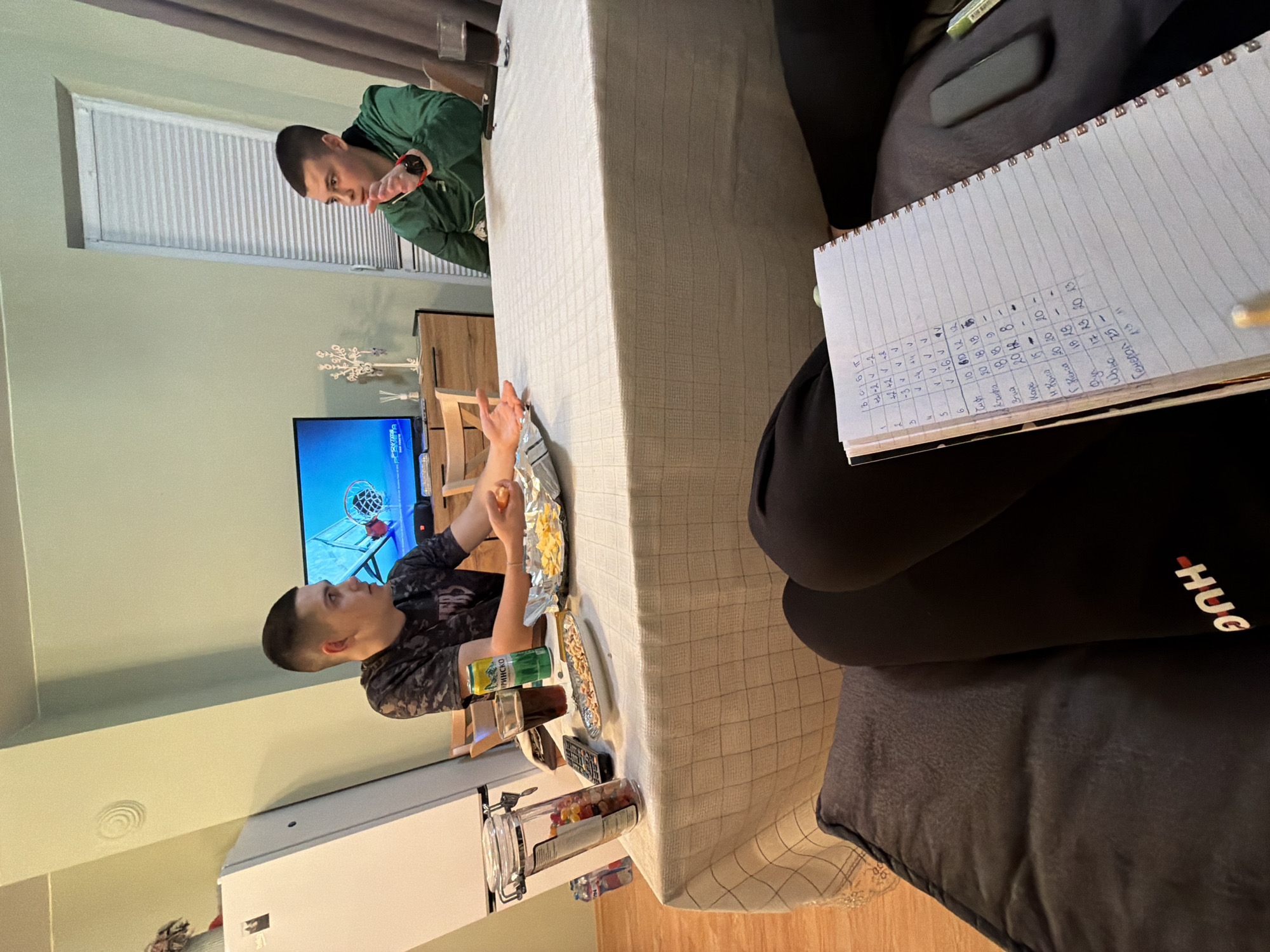 The image depicts an indoor scene. Two men are present, with one eating and watching a television displaying a basketball game. The other man stands near the edge of a table covered in a checkered tablecloth, looking on. Various snacks and beverages are scattered on the table. A notebook with handwritten notes, possibly scores, lies on a dark-colored surface next to the table. The room has light-colored walls, a white refrigerator, and blinds on the window. The location and its relevance to Kavarna, Bulgaria, are not evident in the provided image.