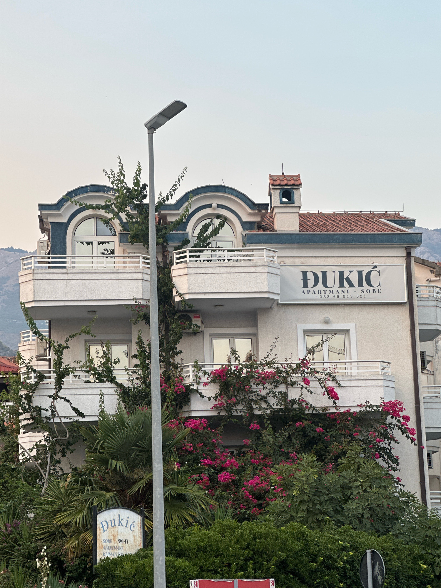 A multi-story building with balconies and arched windows is visible in Budva, Montenegro. The building has a sign that reads "ĐUKIĆ APARTMANI - SOBE +382 69 513 521". The building is adorned with lush green vines and vibrant pink flowers. A street light stands in front of the building. In the foreground, a smaller sign reads "Đukić SOBE WI-FI APARTMANI KLIMATIČNI". The sky is overcast and a faint mountain range can be seen in the background. The overall scene depicts a residential building with a focus on its amenities for guests, set against a backdrop of natural beauty.