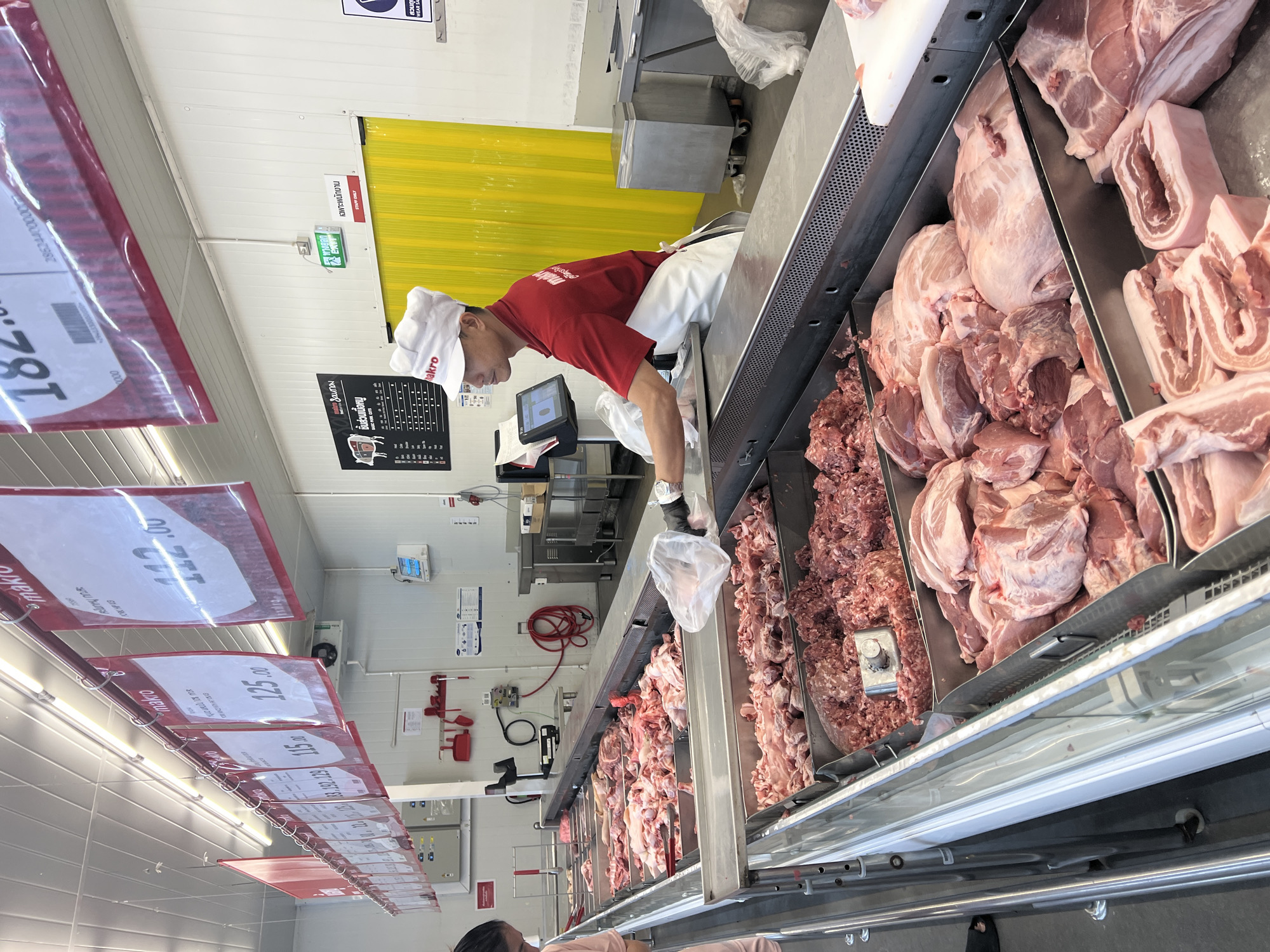 The image depicts a butcher in a Makro store in Baan Hua Thanon, Thailand, preparing meat at a display counter. The butcher, wearing a red shirt, white apron and hat with the "Makro" logo, is using gloved hands to handle meat and place it in a plastic bag. The counter showcases various cuts of pork, including ground meat, chunks of pork shoulder, and pork belly. Overhead, price tags hang from the ceiling indicating the prices of the various cuts. The store interior is utilitarian with white walls and a yellow plastic curtain partially visible. Emergency exit signage is visible above the butcher.