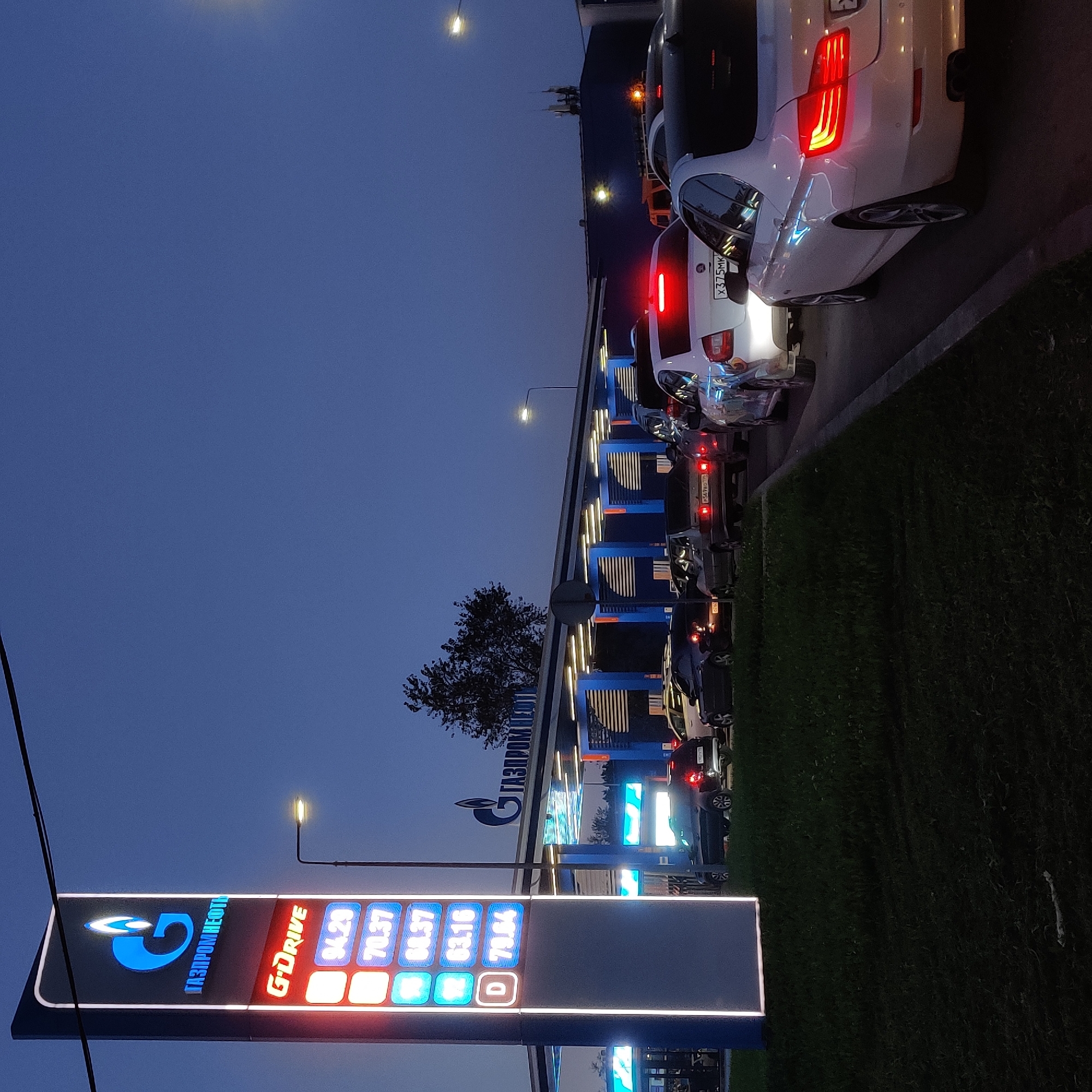 The image depicts a gas station at dusk, with a prominent illuminated sign displaying the "GAZPROMNEFT" logo and fuel prices. The prices shown are 94.29, 70.37, 68.37, 63.16, and 73.84. A queue of cars is visible, waiting to refuel. The gas station has blue accents and overhead lighting. The environment appears to be a roadside in Yekaterinburg, Russia, as indicated by the license plates on the vehicles which conform to Russian standards. The sky is a deep blue, characteristic of twilight. No people are clearly discernible in the image. The overall scene conveys a sense of routine at a busy urban gas station during evening hours.