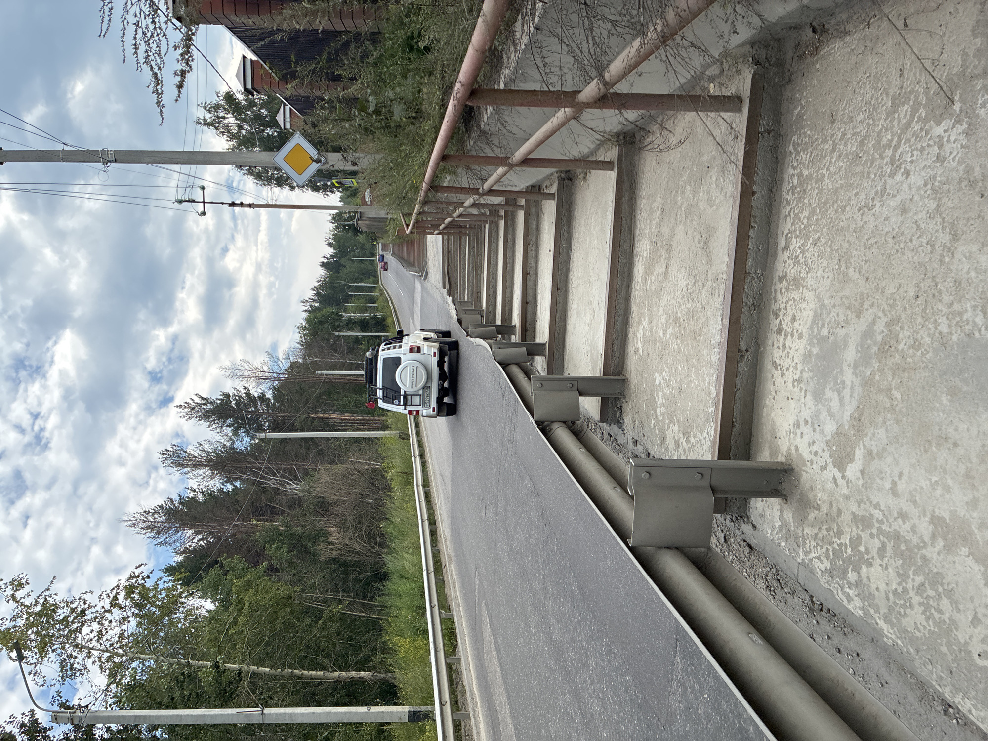 An outdoor daytime scene shows a road on the left and a concrete staircase on the right. The road is asphalt and curves uphill to the left. A white SUV is driving away from the camera on the road. The staircase is made of concrete and has a rusty metal handrail on the right. Trees and vegetation line the road and the top of the staircase. Power lines and poles are visible in the background. A diamond-shaped yellow traffic sign is mounted on a pole near the top of the staircase. The location is identified as Markova, Russia.