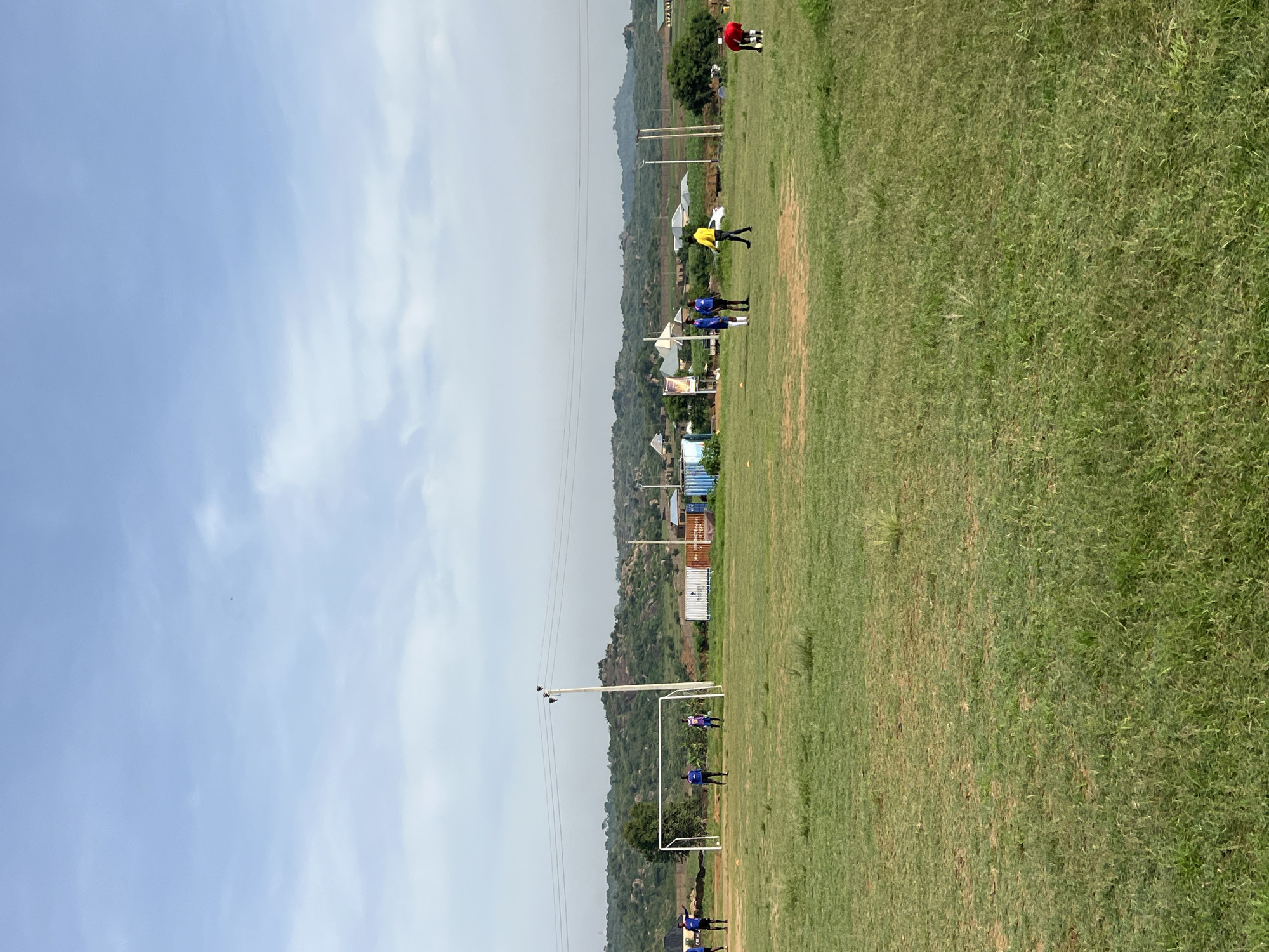 The image depicts an outdoor sports field in Jos, Nigeria, with a clear sky and scattered clouds overhead. The field itself is covered in green grass, with some patches of exposed earth. In the foreground, the grass is well-maintained, while further back, it appears a bit sparser.
Several individuals are present on the field, seemingly engaged in a soccer practice. On the left side, three people in blue uniforms are visible, with one appearing to be interacting with a soccer goalpost. To the right of the goalpost, another individual in a purple and white uniform is standing. Further to the right, a person in a yellow shirt and dark pants is walking, and near them, a person in a red shirt and dark shorts is bending down with a soccer ball.
In the background, various structures are visible, including buildings that appear to be shipping containers, some with signage. A tall utility pole with power lines stretches across the middle ground. Beyond the field and structures, a hilly landscape with scattered vegetation and trees forms the horizon, under the vast expanse of the sky. The overall impression is a scene of activity on a sports field in a semi-arid environment.