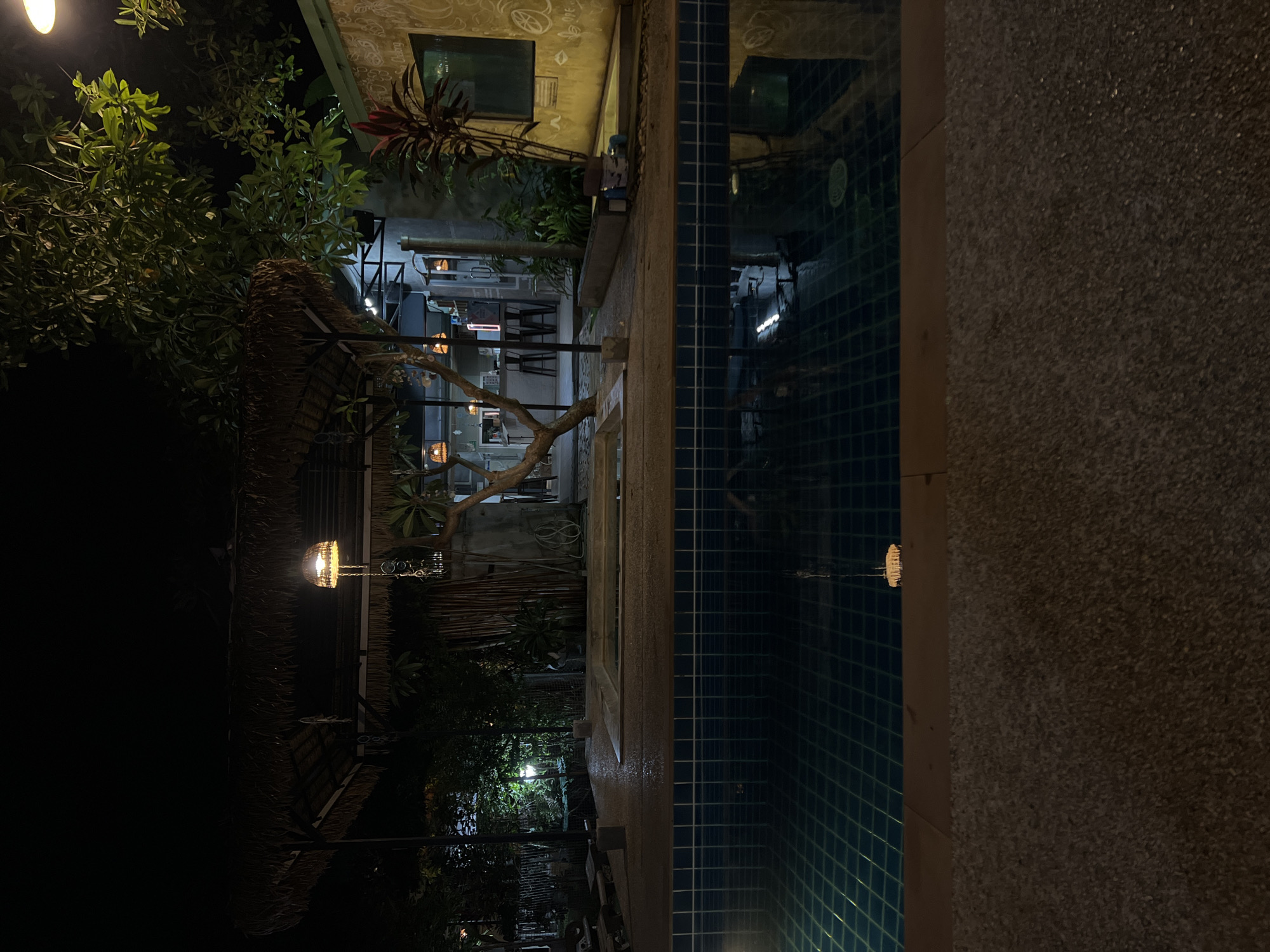 At night, a swimming pool with dark blue tiles is in the foreground. Beyond the pool is a stone patio and a thatched roof structure with hanging lights. A building with a rough, light-colored wall is visible to the right, with a window and some graffiti. Trees and foliage surround the area, and the overall scene is dimly lit by artificial lights. There are no people visible in the image.