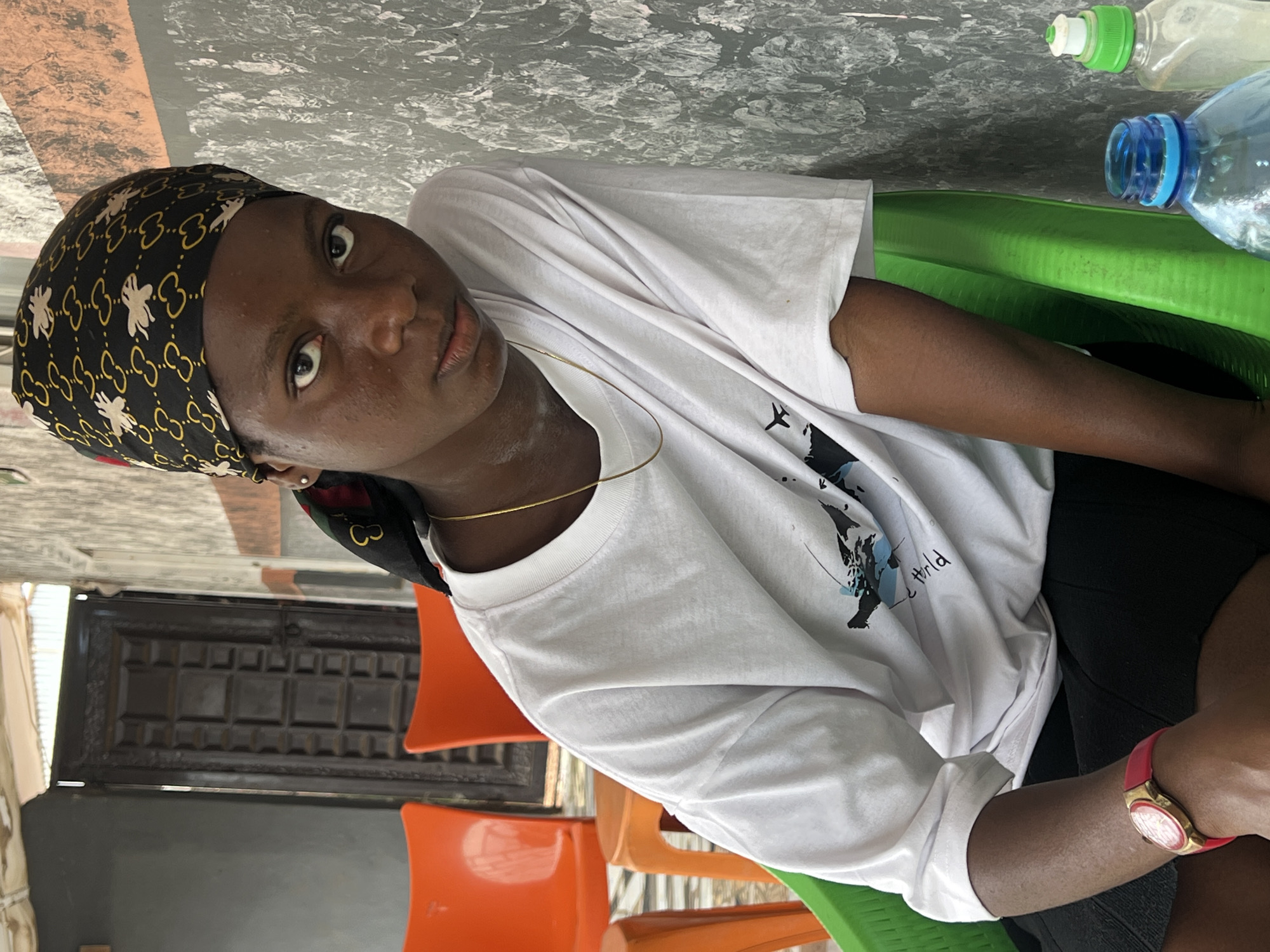 A young Black person with a headscarf patterned with interlocking golden shapes and white bee-like figures, and a white t-shirt with a graphic design, sits in a bright green plastic chair. Their skin is brown, and they have large, dark eyes. A thin gold chain is visible around their neck. A red-strapped watch with a gold face is on their left wrist. They are looking directly at the camera.
The background consists of an abstract, textured gray wall to the right and a dark wooden door with a grid pattern on the left. Two bright orange plastic chairs are visible behind the person, slightly out of focus. A bottle of clear liquid with a blue cap and another bottle with a green cap and white top are on a surface in the foreground. The overall scene suggests an outdoor setting, possibly a shaded patio or veranda in Nkpor, Nigeria. The lighting is somewhat diffused, indicating an overcast day or shaded area.