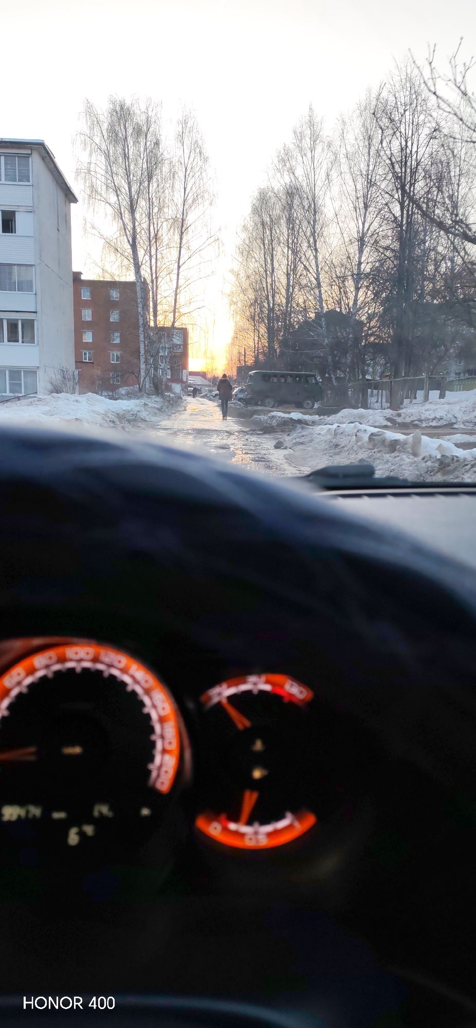 The image captures an outdoor scene in Noviy, Russia, viewed from inside a vehicle during twilight hours. The foreground is occupied by the blurred upper portion of a dark steering wheel and dashboard. Visible dashboard elements include an orange-illuminated speedometer and tachometer, with a digital display showing "6°" (likely temperature) and "9947" (possibly mileage or time).
Beyond the vehicle's windshield, the environment is characterized by thawing snow conditions. A wet, partially cleared dirt or asphalt road, reflecting the sky, extends into the distance. Significant snowbanks line both sides of the road, with puddles and slush present on the surface. Bare-branched deciduous trees, silhouetted against a bright, hazy sky, line the road and are interspersed among buildings.
On the left, a multi-story light-colored residential building with several windows is visible, partially obscured by the vehicle's interior. Behind it, a darker, brick-colored multi-story building stands. In the middle ground, a lone individual in a dark jacket walks away from the viewer along the wet road toward the distant horizon. The sun, a bright orange glow, is low in the sky, illuminating the distant end of the road and casting a warm hue. To the right, a dark green utility van, possibly a UAZ-452, is parked facing away from the camera, partially obscured by snowdrifts. Additional indistinct figures and objects are visible further down the road.