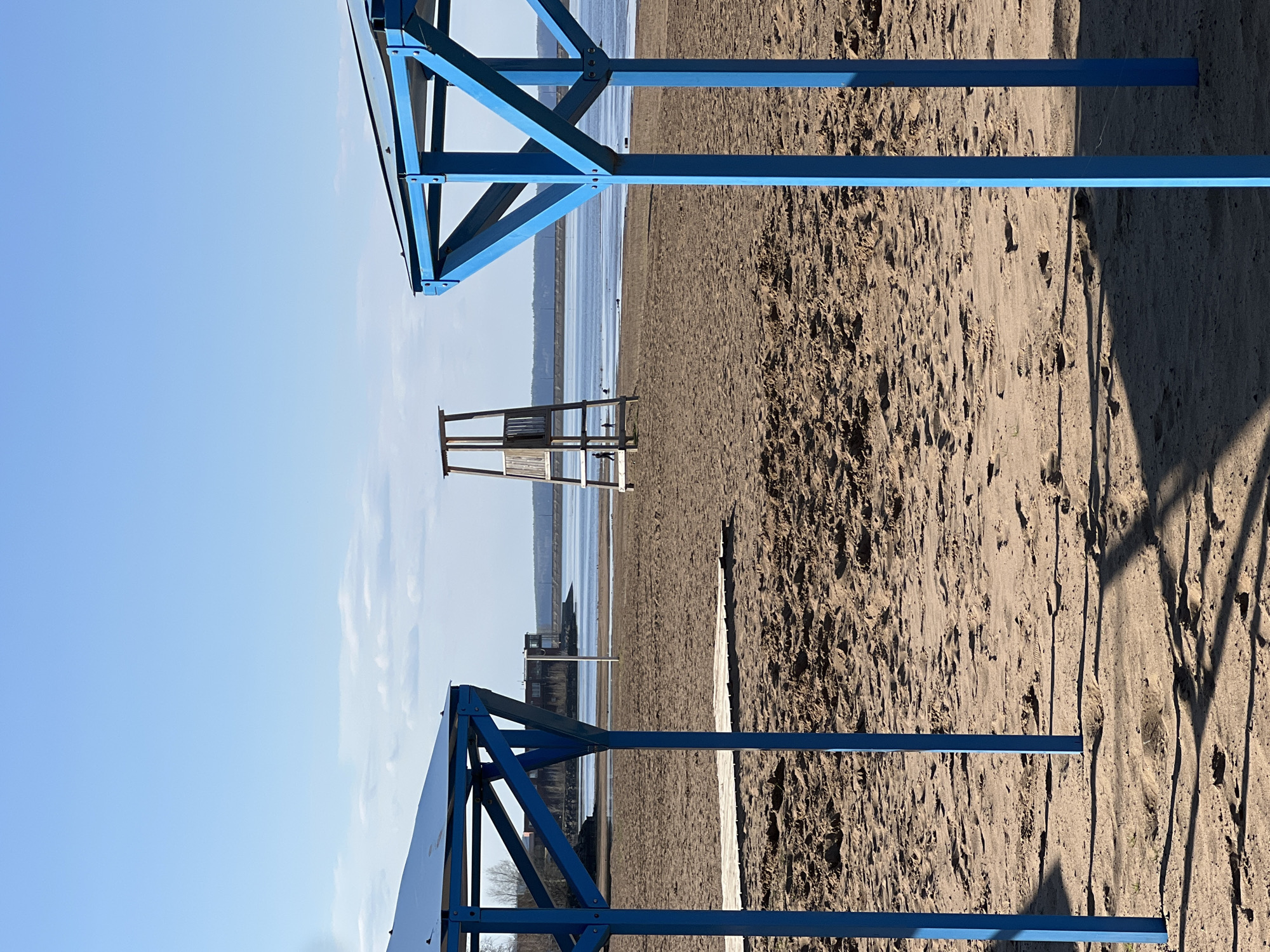 The image shows a beach scene in Chaikovsky, Russia. The foreground is dominated by a sandy beach, with patterns and textures indicating footprints and natural sand formations. A blue, metal framework structure stands on either the left or right edge of the image.
In the midground, there is a lifeguard chair. Further in the background is a body of water, with a hazy horizon. Along the water's edge, there are buildings or structures, suggesting a populated area.
The sky is clear blue, indicating a sunny day. The overall scene is still and quiet, possibly capturing an off-season moment at the beach.