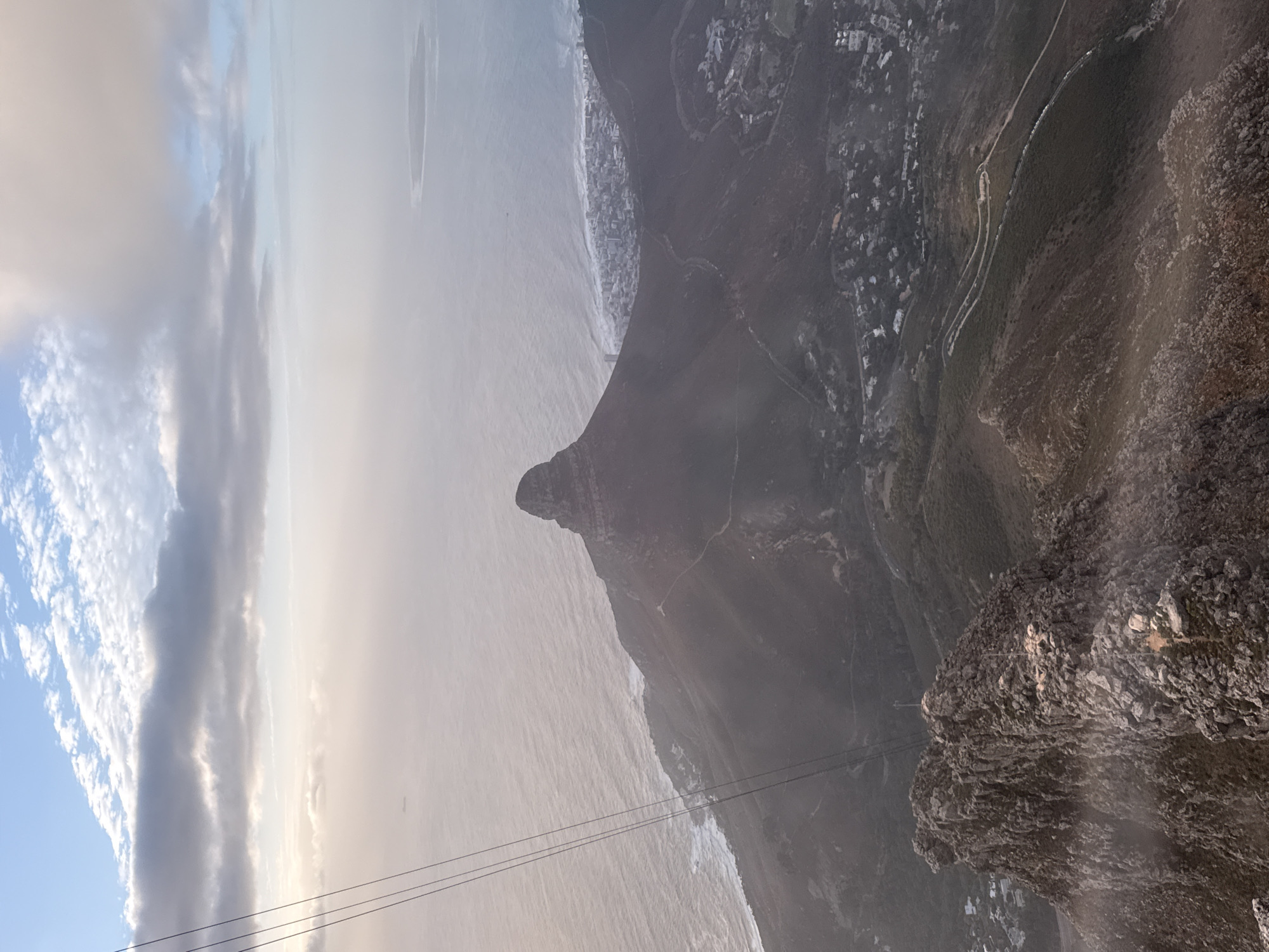 The image captures a landscape near Cape Town, South Africa. It appears to be taken from an elevated viewpoint, possibly Table Mountain. The prominent feature is Lion's Head, a peak jutting out towards the Atlantic Ocean, separated by a narrow strip of land from the coastal city. The ocean meets the shoreline, and a hazy atmosphere obscures the distant horizon. Cable car lines are visible in the lower portion of the image. The foreground shows rocky terrain and vegetation. A road with visible buildings is along a mountainside. Clouds and the sky are in the upper left portion of the image.
