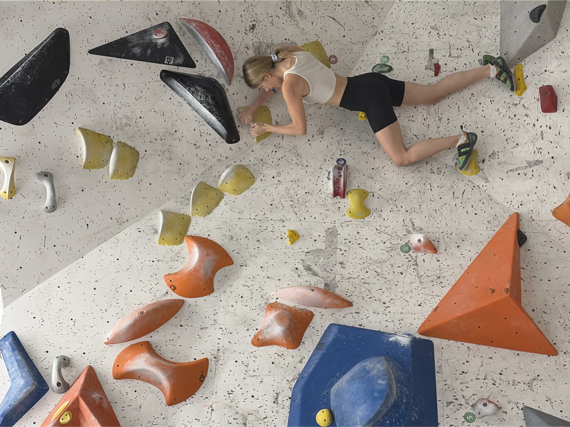 The image shows a young woman climbing on a brightly lit indoor climbing wall in Khimki, Russia. The wall is covered with numerous climbing holds of various shapes, sizes, and colors, including orange, yellow, black, blue, and red. The woman, wearing a white sports bra and black athletic shorts, is positioned midway up the wall. Her right hand is gripping a yellow hold, and her left hand is reaching for another yellow hold above her. Her legs are bent, with her right foot on a yellow hold and her left foot on a yellow hold below her. The wall has a textured, speckled surface with visible chalk marks from previous climbers. In the background, more climbing holds and sections of the wall are visible. The overall environment suggests a dedicated indoor climbing facility.