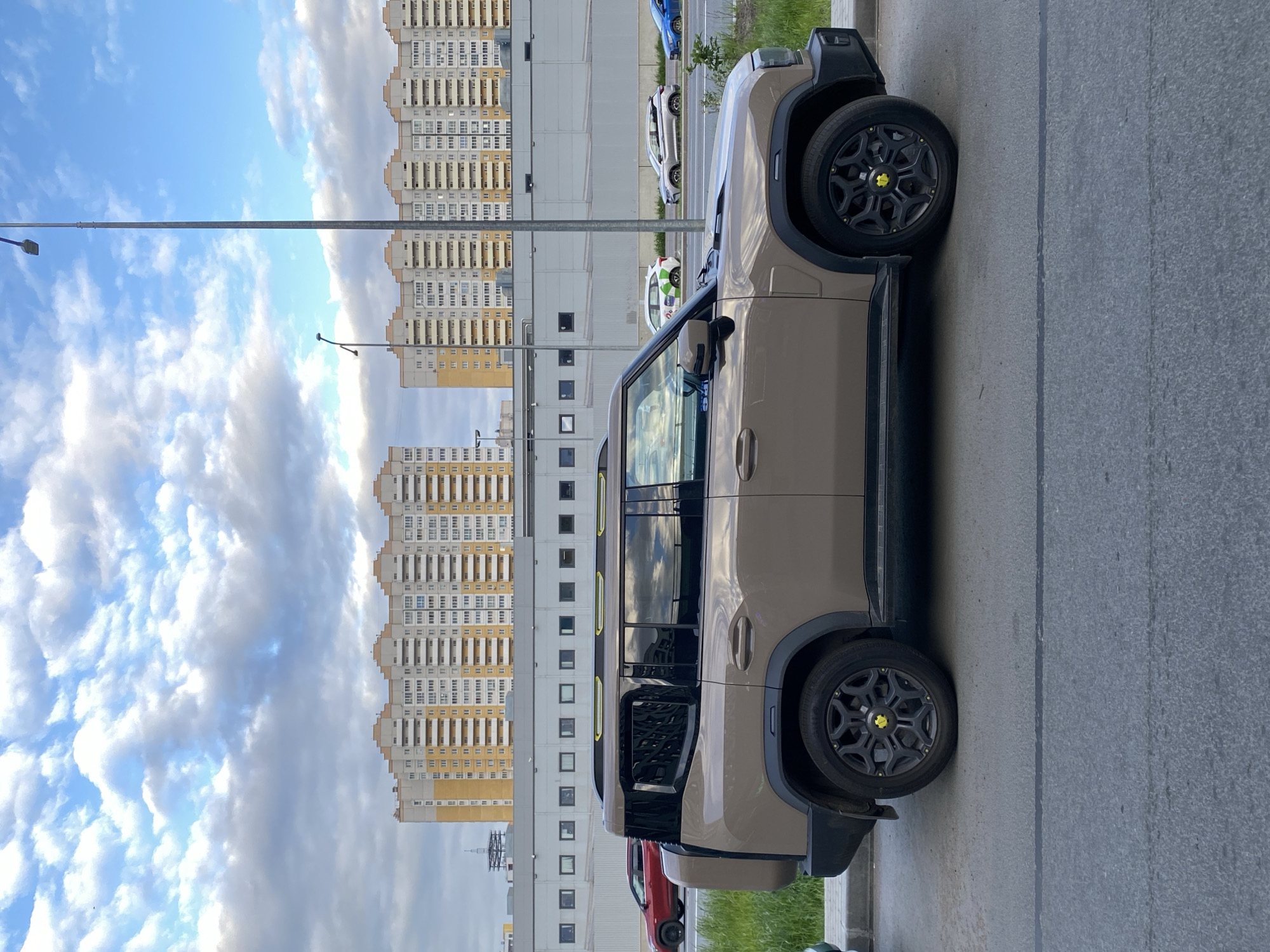 The image depicts a beige Rivian R1S electric SUV parked on a paved surface, likely a street or parking lot. The vehicle has black wheels with yellow accents. In the background, there are tall residential buildings with a repetitive window pattern, indicative of a modern urban area. The sky is partially cloudy with patches of blue. Other parked cars, including a red one, are visible. The presence of modern high-rise buildings and the type of vehicles suggest an urban environment, and the presence of Russian architectural styles indicates the location of Saint Petersburg, Russia. The presence of electric vehicle suggests a certain degree of wealth.