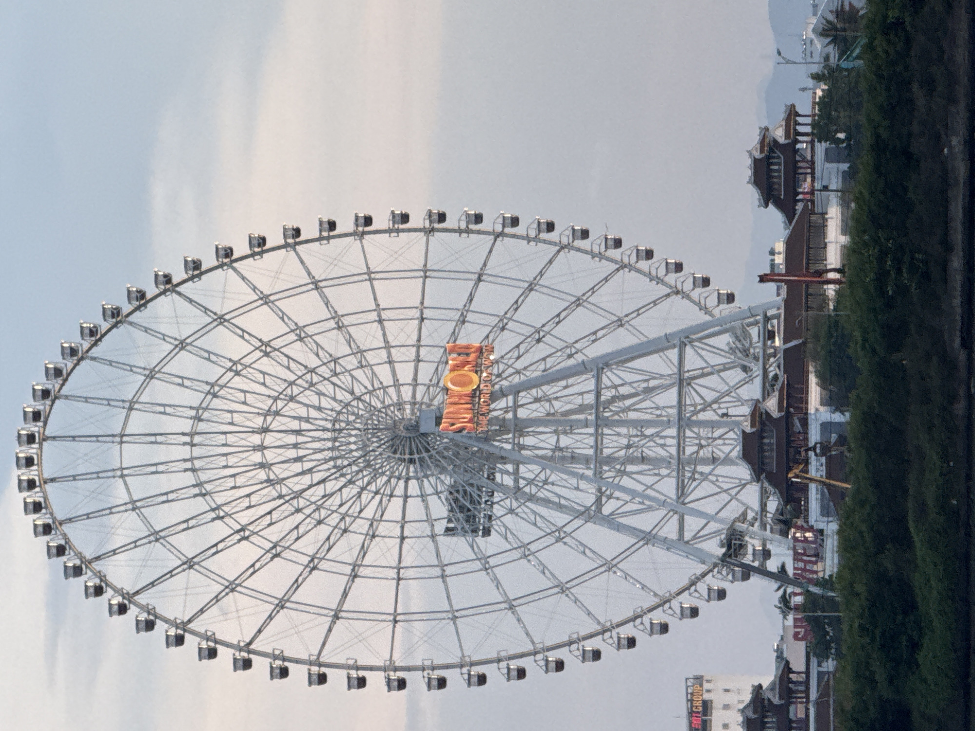 The image shows a large ferris wheel in Da Nang, Vietnam, under a pale, overcast sky. The ferris wheel dominates the frame, with its intricate metal structure and numerous passenger cars visible. Orange lettering on the ferris wheel spells out "THE WORLD'S BIGGEST". In the background, traditional Vietnamese-style buildings with tiered roofs are visible, nestled amongst lush green trees. Further in the distance, hills can be seen. The scene appears to be set in an amusement park or a recreational area. There is no explicit indication of people, nor is any specific activity or event taking place. The time of day appears to be daytime, possibly late afternoon or early evening, given the soft lighting.
