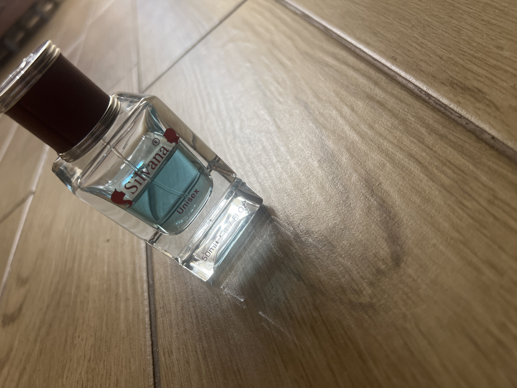 A clear glass bottle of "Silvana" brand unisex perfume, containing a light blue liquid, rests on a light brown wooden surface. The bottle has a dark red cap. Text on the bottle reads "Silvana", "Unisex", and "50ml e 1.7 Fl.Oz.". The scene is illuminated by overhead lighting, creating a shadow to the left of the bottle. The wooden surface appears to be made of planks. The location is the city of Chernorechenskoye, Russia.