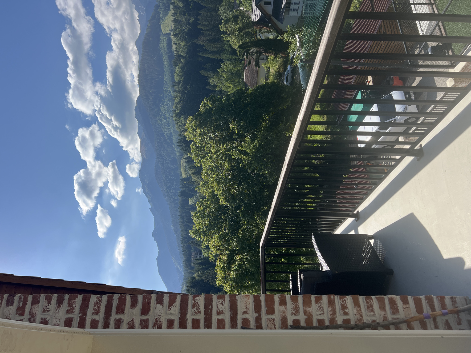 A clear, sunny day is depicted from a balcony overlooking a mountain landscape. The foreground shows the edge of a brick wall and a railing made of dark metal. A portion of the balcony floor is visible, with shadows cast from unseen objects. Beyond the railing, a variety of trees dominate the midground, with a few houses nestled among them. In the background, a range of layered mountains stretches across the horizon under a blue sky dotted with fluffy white clouds. The overall scene is one of natural beauty and tranquility.