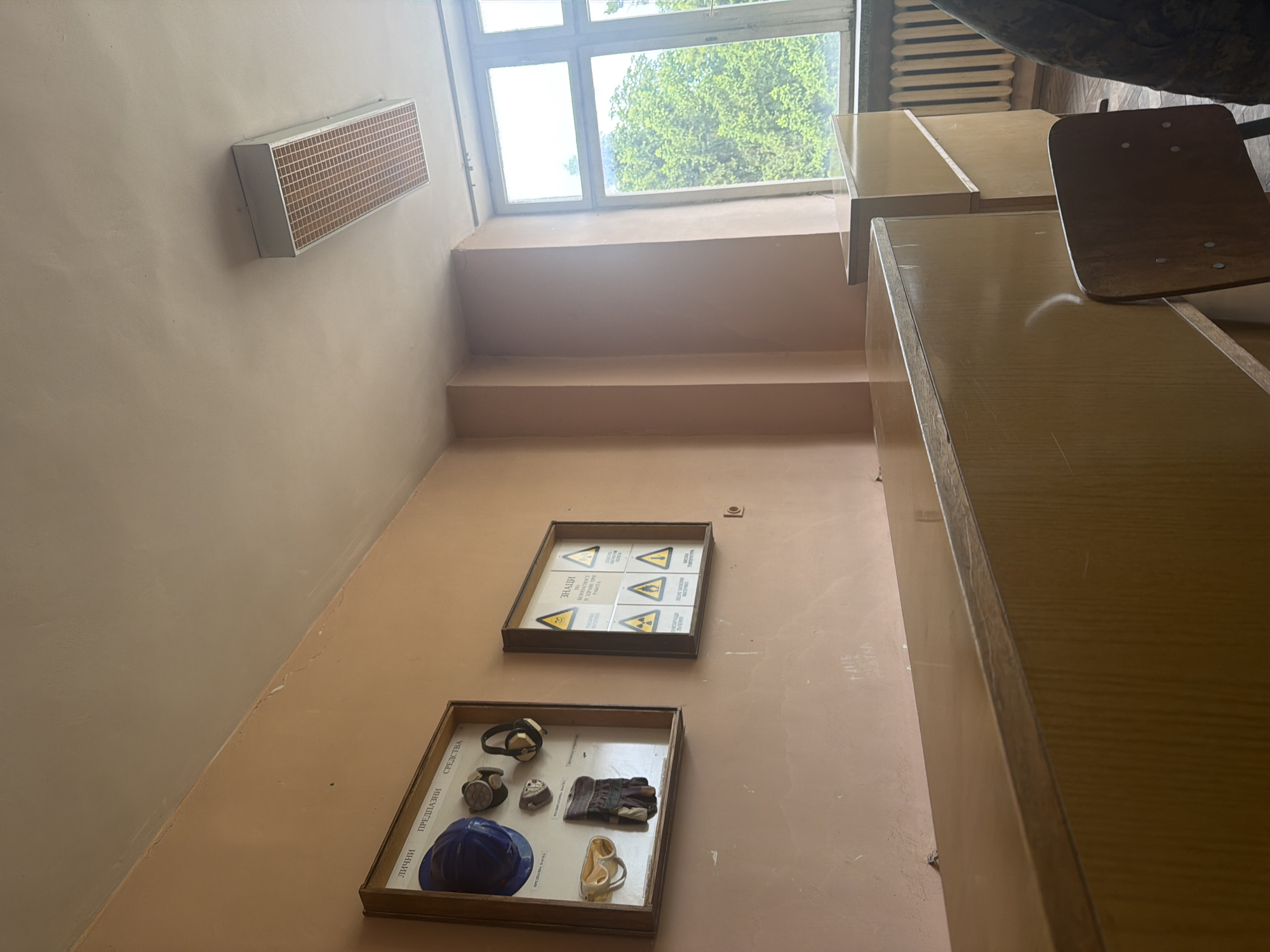The image depicts an interior space, likely a building in Dolna Mitropolia, Bulgaria. The walls are peach-colored, and there is a window with a view of green trees outside. On the wall are two framed displays: one appears to contain safety equipment, including a blue helmet, gloves, and respirators, while the other contains radiation hazard symbols. A rectangular ventilation unit is mounted high on the wall. Desks and a chair occupy the right side of the frame. The perspective is from a low angle, looking upwards towards the window.