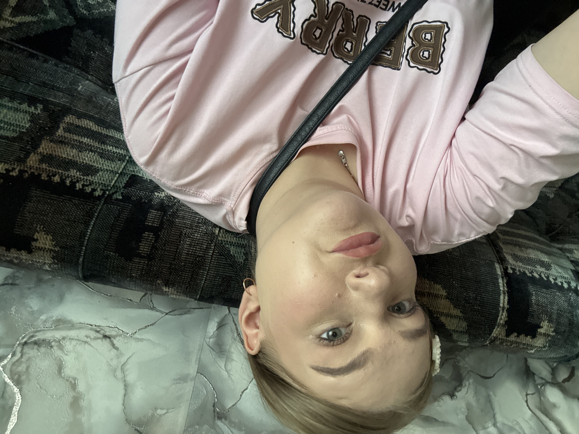 A woman with fair skin, light brown hair, and blue-gray eyes is lying on a surface that appears to be a combination of a patterned dark fabric and a light-colored surface with a marble-like pattern. She is wearing a light pink long-sleeved shirt with brown lettering. A black strap crosses her chest. The location is unknown, and there is no information to suggest the location is in the city of Ольховка, Russia.