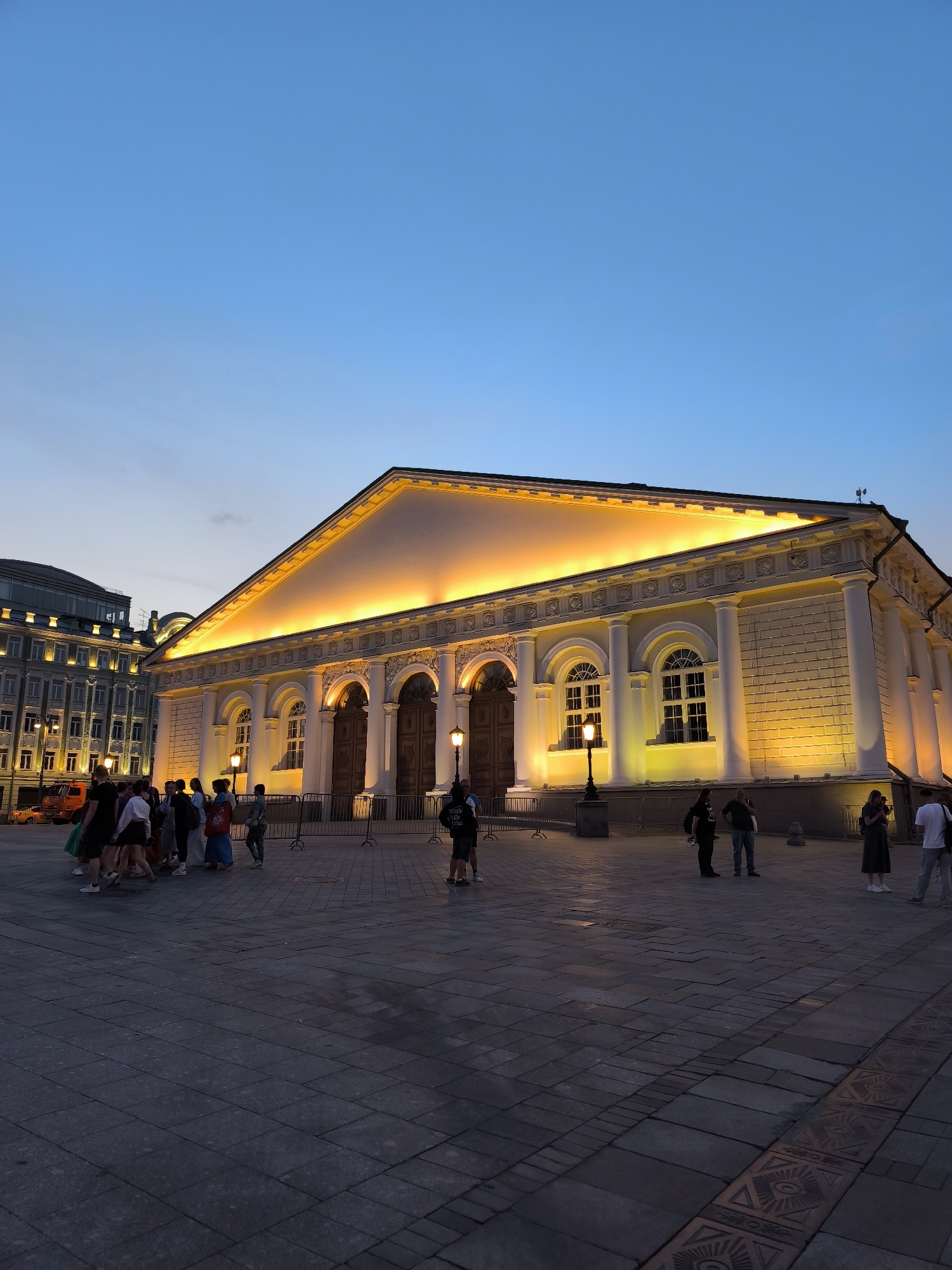 The image captures an exterior view of the State Tretyakov Gallery in Moscow, Russia, at dusk. The building, an imposing neoclassical structure, is illuminated by warm yellow lights that highlight its architectural details, including arched doorways, columns, and ornate friezes. The sky above is a clear gradient of deep blue to lighter blue, indicating the twilight hour.
In the foreground, a group of people are gathered on a wide, paved plaza. They appear to be tourists or visitors, some pointing and gesturing, engaged in conversation. Their clothing is casual and summery. The paving stones of the plaza are made of large, square tiles, with some decorative patterned tiles visible in the foreground.
To the left of the Tretyakov Gallery, another building with numerous illuminated windows is visible, suggesting a surrounding urban environment. A bright orange vehicle, possibly a fire truck, is parked near this building. Streetlights stand on the plaza, casting pools of light. The overall atmosphere is one of a serene evening in a significant cultural landmark.
