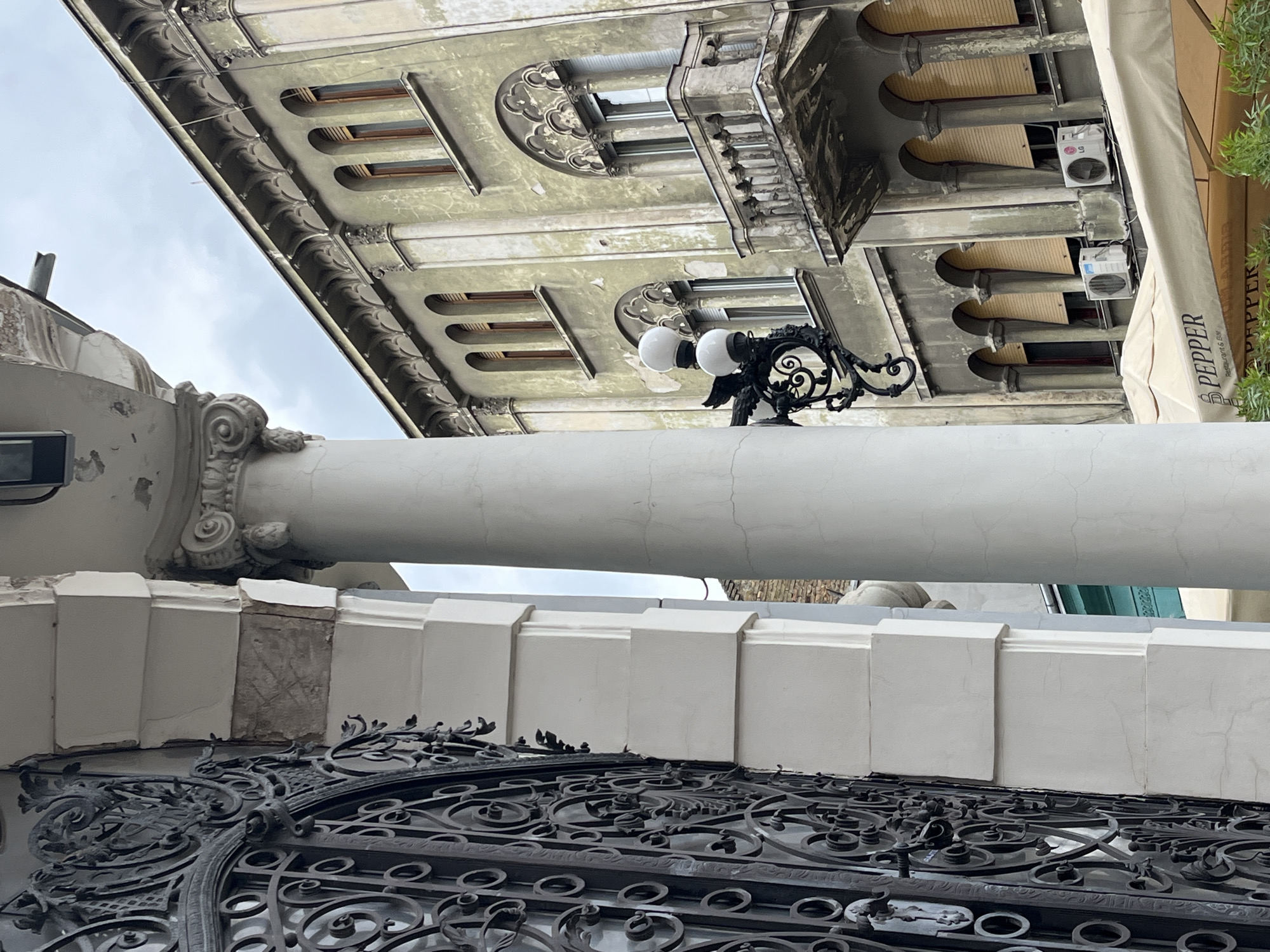 A low-angle shot showcases architecture in Novi Sad, Serbia. A black ornate gate, located at the bottom of the frame, leads to a building. The building has a white column that supports an outdoor light fixture with two round white globes. Above the light fixture, the building's facade includes windows with arched detailing and horizontal bars, air conditioning units, and a brown awning with the word "PEPPER" printed vertically. The sky is overcast and visible in the upper portion of the frame.