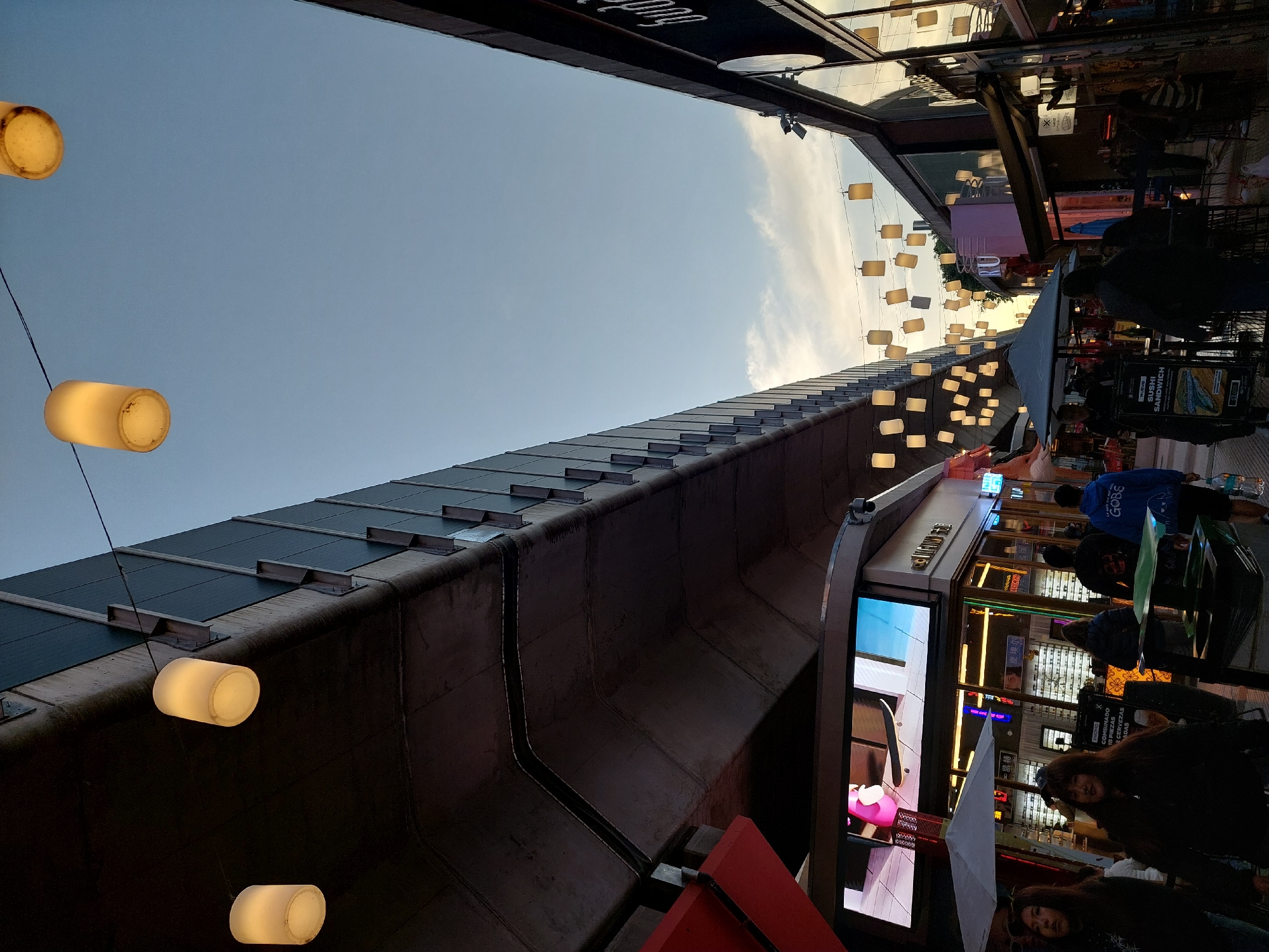 The image shows a street scene, possibly in Buenos Aires, Argentina, based on the street style. The view is looking upwards at the buildings and sky. There are cylindrical lanterns hanging above the street. Some people are visible, seemingly dining outdoors. Buildings flank the street with shops or restaurants. A large structure, possibly a bridge or building overhang, is a prominent feature above.
