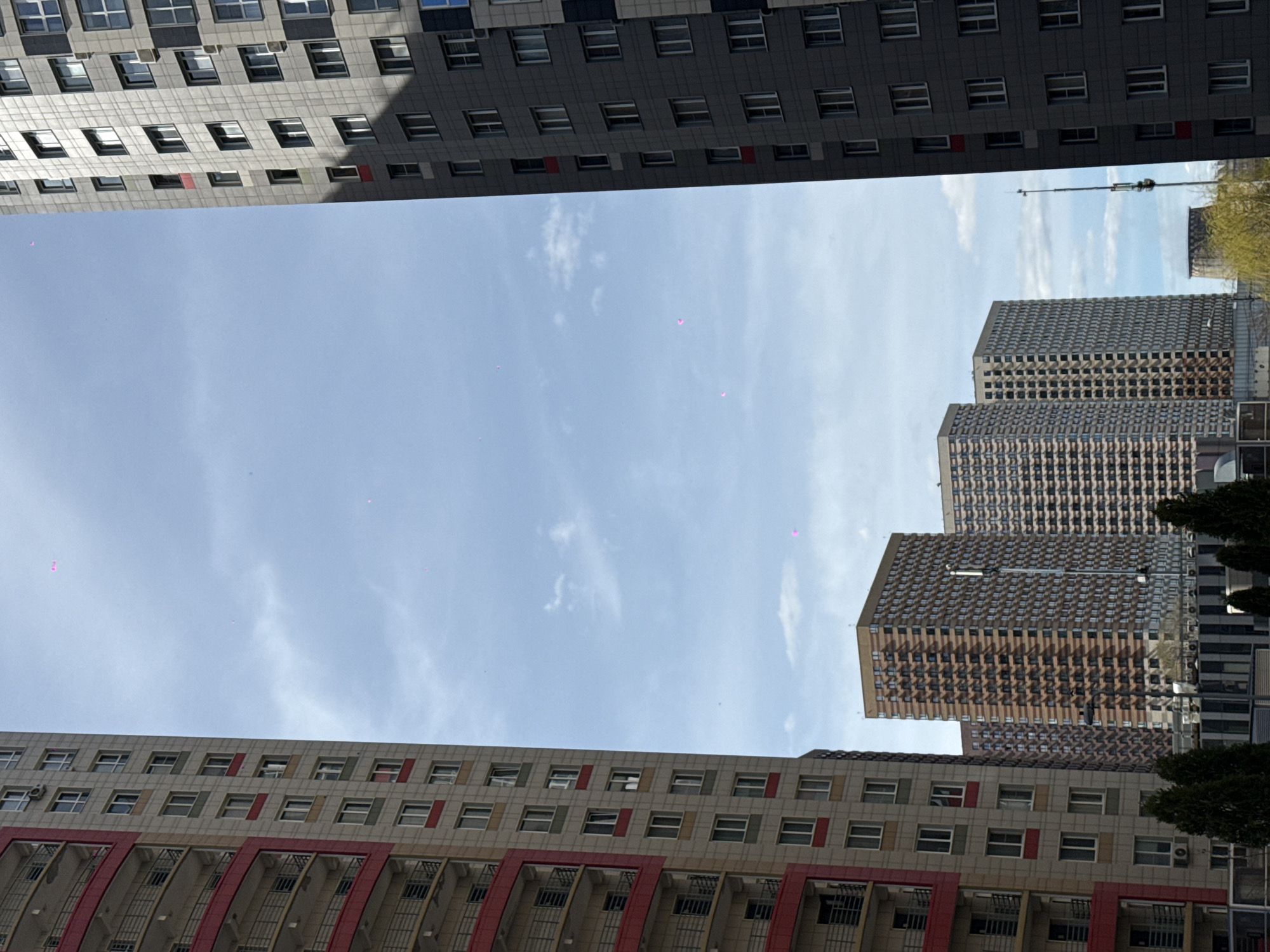 The image shows a view looking up between buildings, possibly in Khimki, Russia, given the description. The sky is light blue with scattered clouds. Buildings frame the view, with a gray apartment building with a grid of windows and red accents at the bottom. A similar gray building with dark windows sits at the top of the frame. In the central right, three taller buildings with a repeating patterned facade stack somewhat staggered. Some green trees are visible on the right side of the photo, partially obscuring the buildings. A communications tower can be seen above and behind the buildings on the right side.