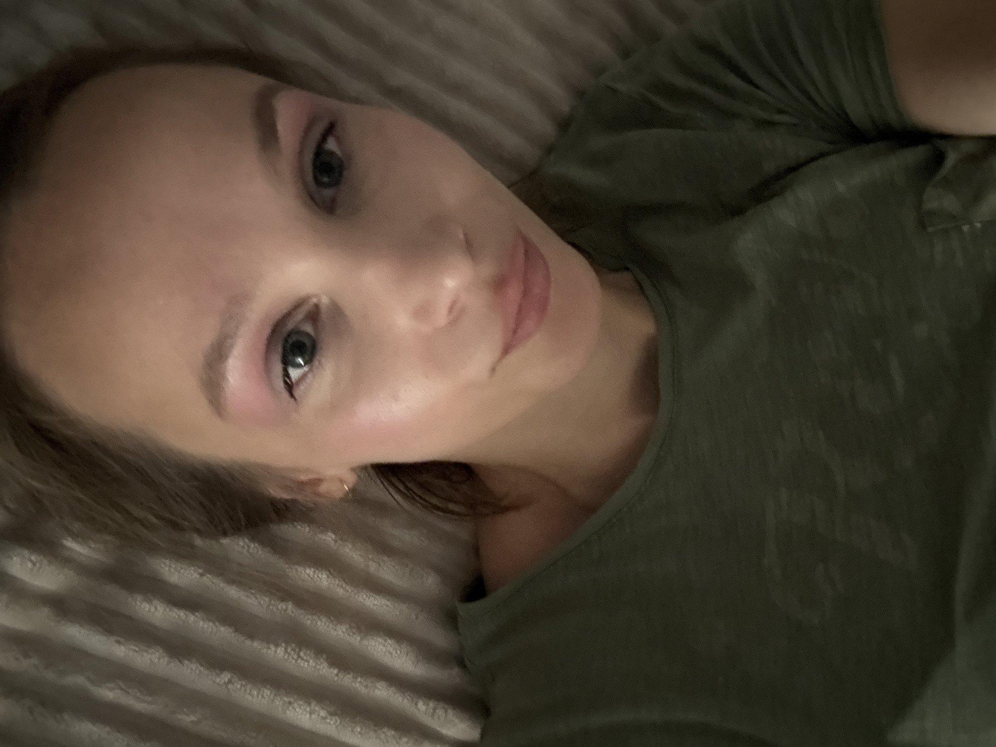 A close-up selfie shows a woman lying down, with her head tilted and eyes looking at the camera. She is wearing a green t-shirt and has pink eyeshadow on. The background appears to be a textured surface, possibly a bed. The image quality is somewhat grainy, and the lighting is dim, suggesting it was taken indoors, likely at night. The woman is positioned centrally in the frame, with her face and upper torso visible. There are no visible indications of the specific location within Überherrn, Germany.