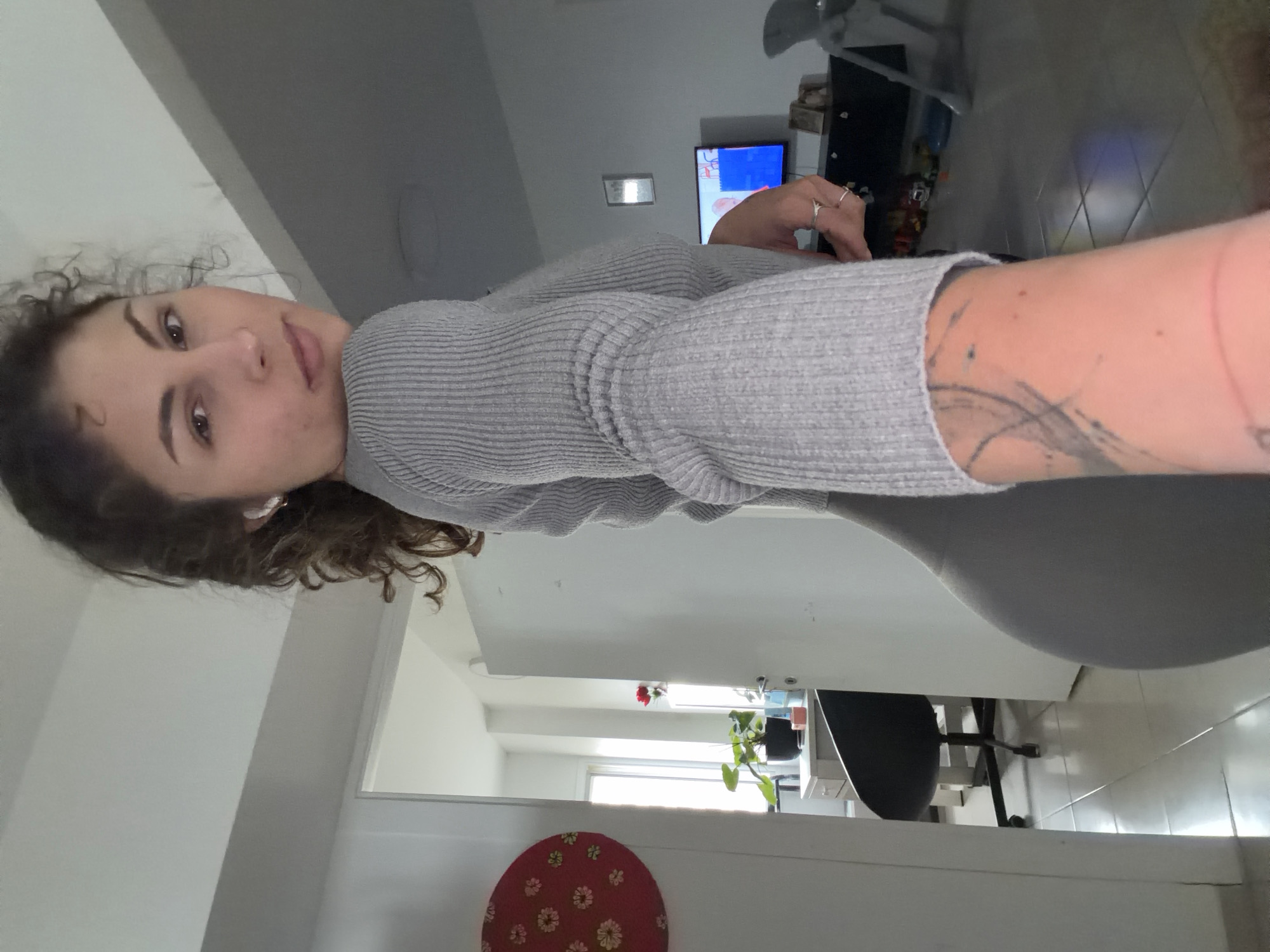 A woman with curly brown hair and fair skin is in a room with a gray shirt. She is likely in Kiryat Bialik, Israel. She has a tattoo on her arm. There is a television in the background. There is a red cushion with a floral pattern on the floor and a green plant on a table. A black office chair is also visible.
