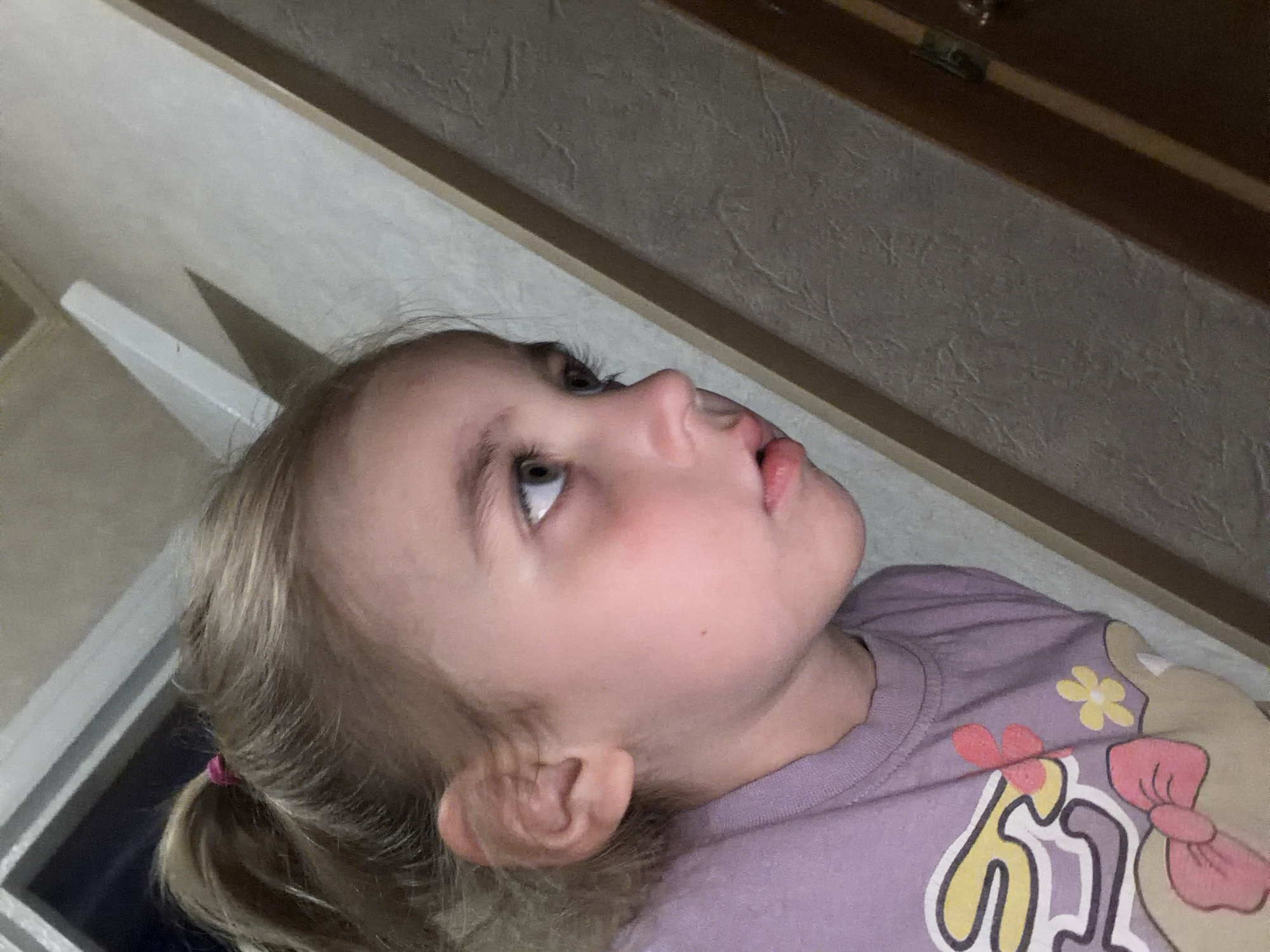 The image shows a close-up, side-profile of a young Caucasian girl. She has blonde hair pulled back in a ponytail with a pink hair tie. Her skin is fair with some pinkish undertones on her cheeks and under her eyes. She is wearing a purple t-shirt with a cartoon graphic featuring a bow and flowers. Her mouth is slightly open, and her eyes are looking to the right of the frame. The background consists of a textured wall with wooden trim and a diagonal beam. The lighting suggests an indoor setting, possibly at night, given the prominent shadow cast by the beam. The image does not contain any information that would allow for the identification of a specific location in Tambov, Russia.