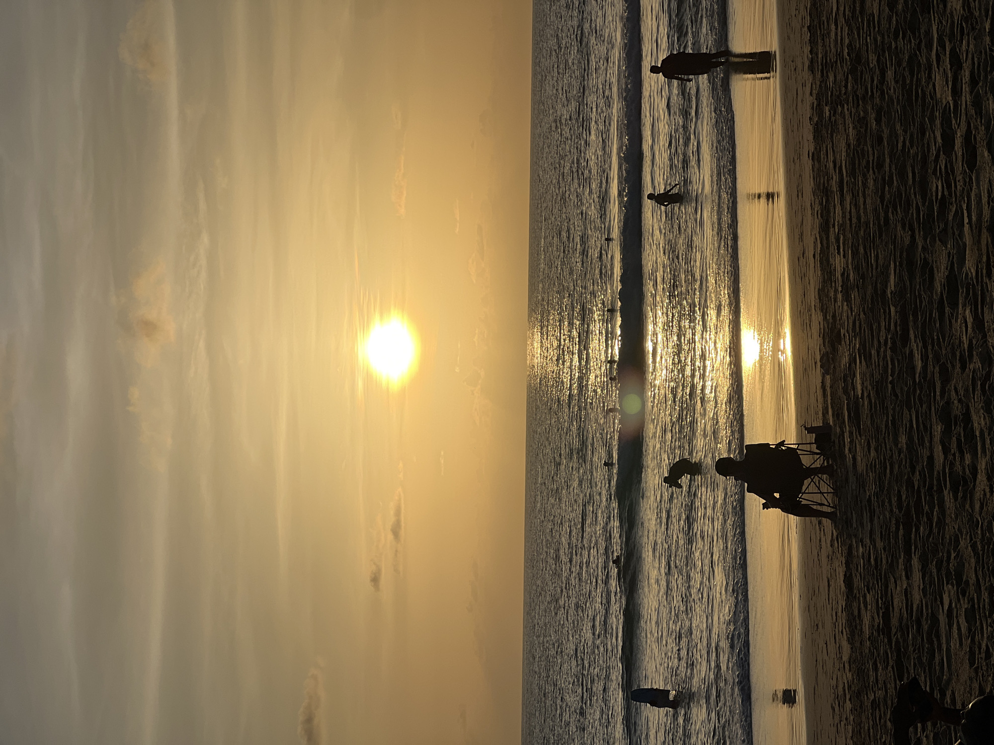 A wide shot captures a beach scene at sunset in Choeng Thale, Thailand. The sun, a bright orb, is positioned slightly right of center in the sky, casting a golden hue across the clouds and the ocean. The water is calm with gentle waves breaking near the shore. Several people are visible in the water, some wading and others appearing to be swimming or surfing.
In the foreground, a person sits in a folding chair on the sandy beach, facing the ocean. The silhouette of the individual is distinct against the bright background. Another person is partially visible in the lower left corner, also silhouetted. The beach sand is textured with footprints and ripples. The overall atmosphere is serene, with the warm light of the setting sun dominating the scene.