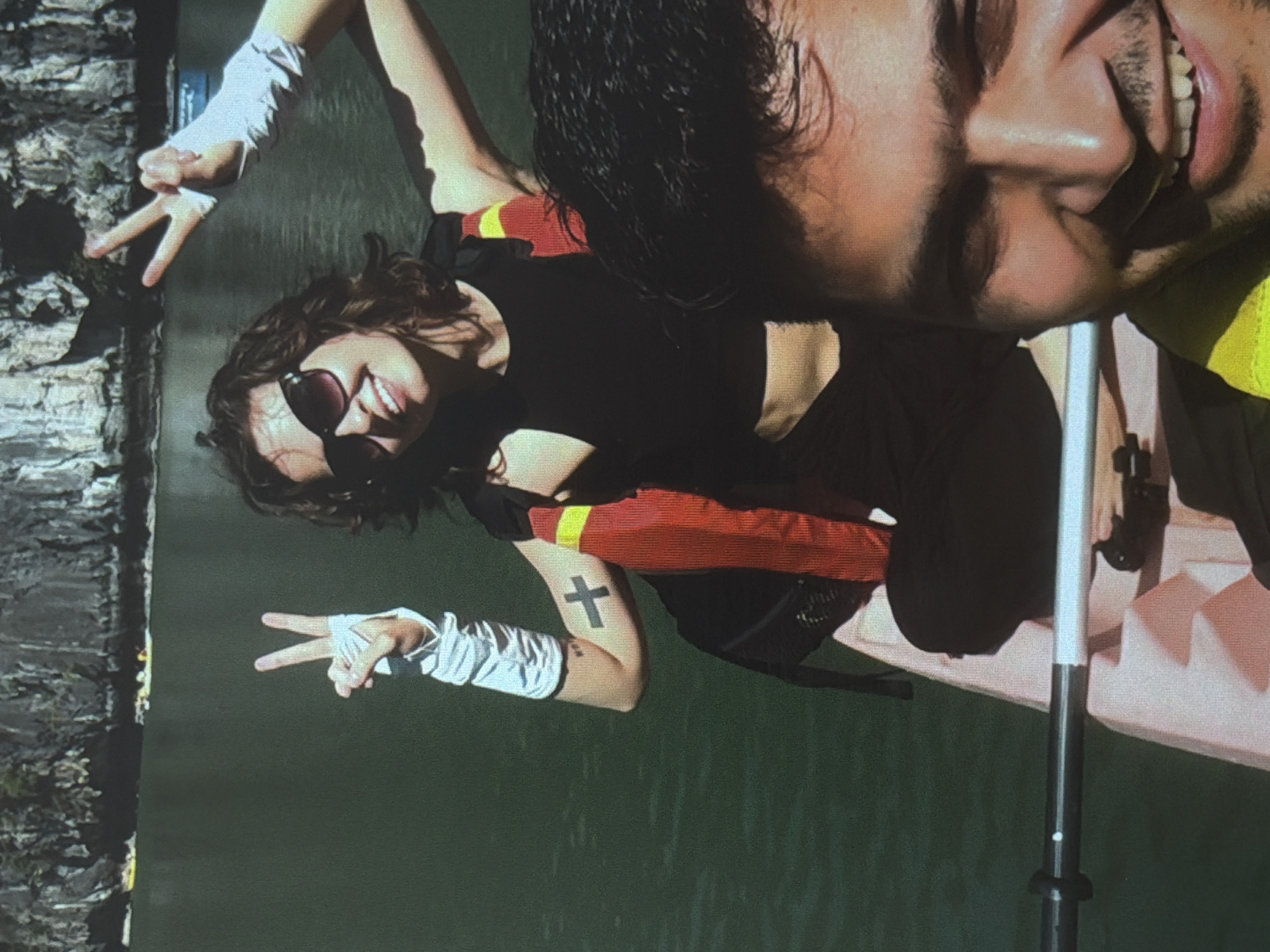An outdoor, brightly lit image captures two individuals enjoying a water activity, likely kayaking or boating, in Đà Nẵng, Vietnam. The scene features calm, dark green water with a textured, dark bank or shoreline visible on the left.
In the foreground, a female with dark, wavy hair is smiling broadly and wearing dark sunglasses. She is dressed in a black crop top and a red and yellow life vest. Her left arm, adorned with a white armband or sleeve, makes a peace sign. A distinct black cross tattoo is visible on her upper left arm.
Partially visible in the upper right is a male, whose face is seen in a close-up, smiling joyfully with teeth showing. He has dark hair and a beard and appears to be wearing a yellow garment, likely a life vest, consistent with the water activity.
The individuals are in a light-colored boat or kayak, with part of a paddle shaft visible. Both subjects exude happiness, indicated by their wide smiles and engaged poses, suggesting a pleasant recreational outing under sunny conditions.