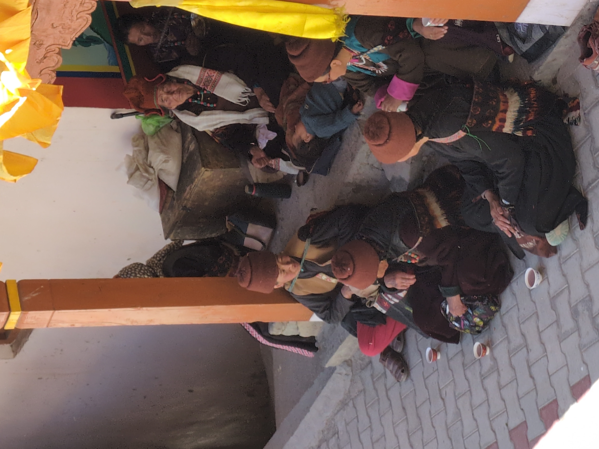 The image depicts a group of people, likely women, gathered outdoors on a paved walkway in Pibiting, India. The individuals are seated on steps and the ground, some appearing to be in conversation. Several people are wearing traditional clothing and headwear, including distinctive red hats. Three small cups containing a dark liquid are placed on the ground in front of some of the seated individuals. The environment suggests a sunny day, with shadows indicating overhead structures. A portion of an orange pillar is visible on the right side of the frame, and yellow fabric hangs above the scene. The background includes a white wall and what appears to be a wooden chest. The overall atmosphere is informal and communal.