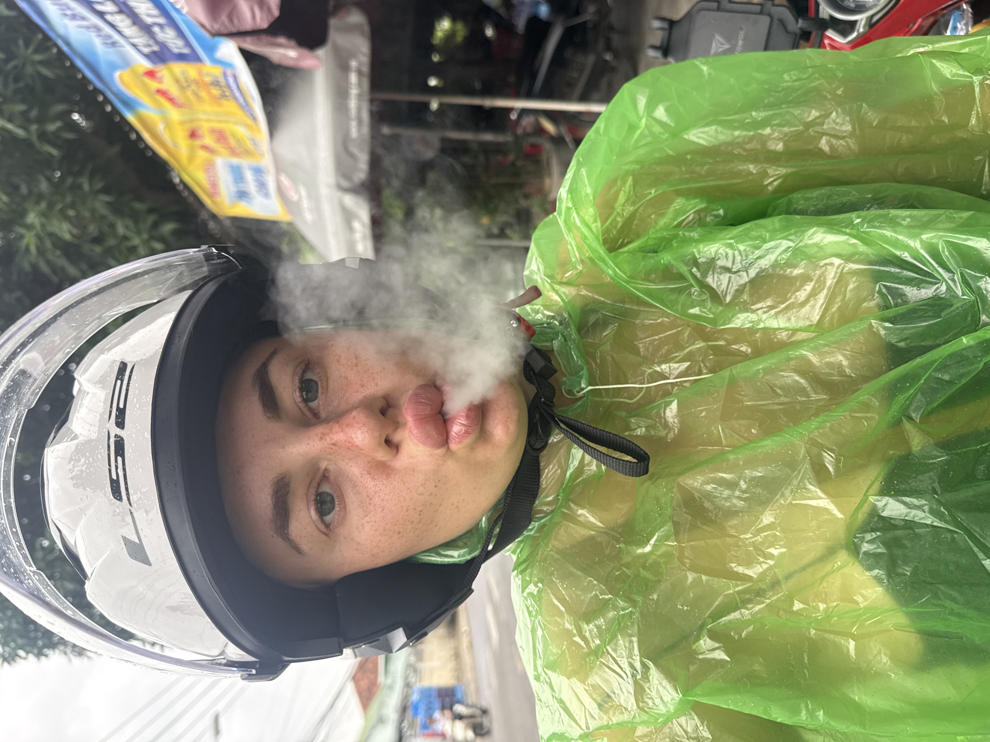 A woman wearing a white helmet and a translucent green poncho is leaning forward, exhaling a plume of vapor. Her lips are pursed, and she has freckles across her nose and cheeks. Behind her, a street scene unfolds in Nha Trang, Vietnam, with trees, power lines, a Red Bull advertisement, and other vehicles visible. The weather appears overcast.