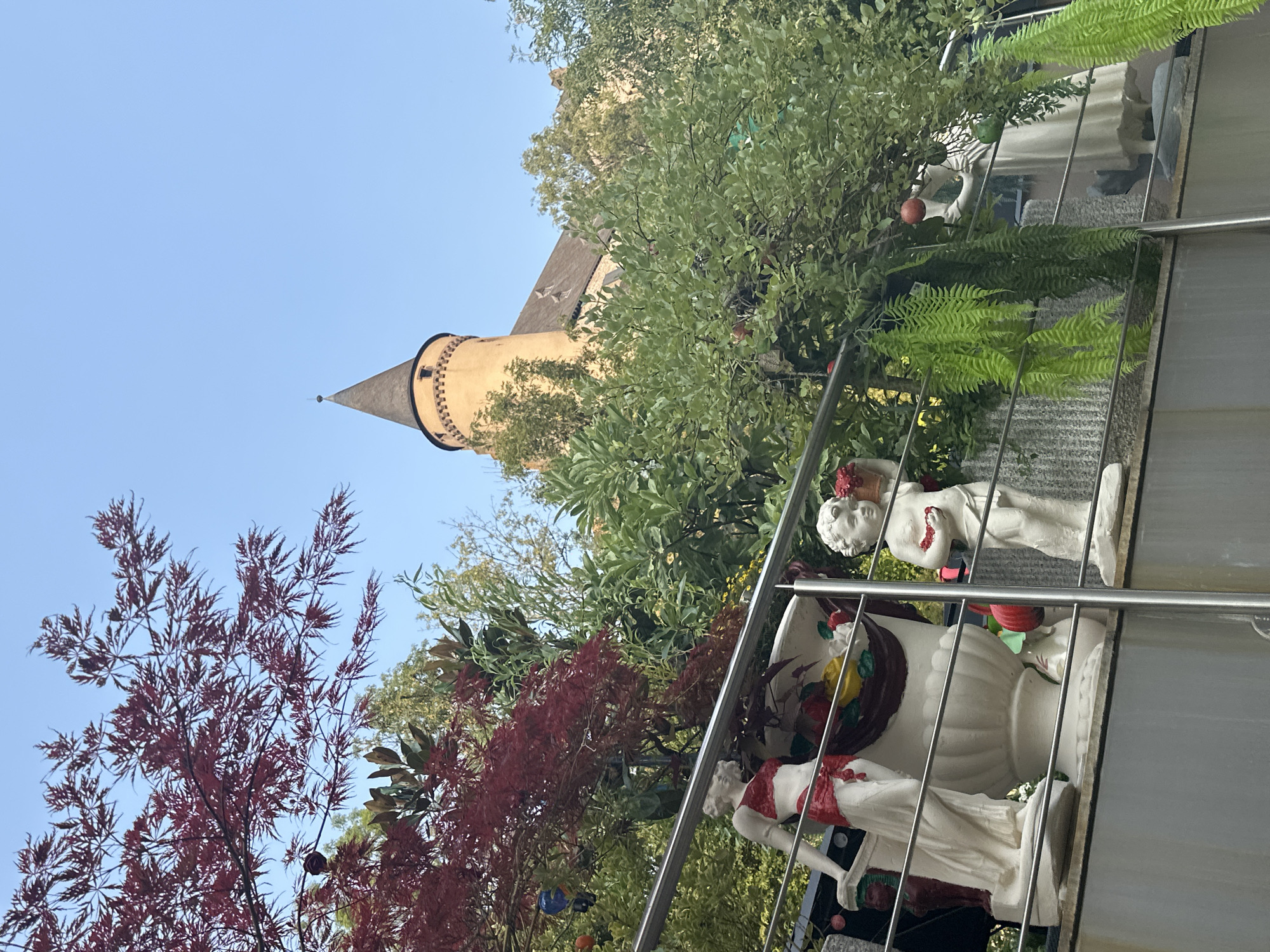 The image captures a view from an elevated position, looking towards a castle tower in Vianden, Luxembourg. In the foreground, a metallic railing runs horizontally across the frame, separating the viewer from decorative sculptures and lush greenery.
On the left, a white statue of a female figure, adorned with red fabric around her waist and chest, stands beside a large, ornate white vase filled with artificial fruits. To the right of the vase is a white statue of a cherubic child holding a red object in its hands and a basket of flowers. Further to the right, another white statue of a female figure in a flowing dress is visible, holding a red apple. Cascading green ferns hang from above, adding to the vibrant foliage that surrounds the statues.
Behind the statues and railing, a dense arrangement of trees and shrubs creates a natural backdrop. Prominent among these is a tree with striking deep red, maple-like leaves, contrasting with the lighter green of other trees.
In the background, the distinctive conical tower of Vianden Castle rises above the foliage, its orange stone structure capped with a dark, pointed roof. The castle appears to be situated on a hill, with a clear, light blue sky overhead. The overall scene suggests a decorative garden or terrace area with a view of the historic castle.