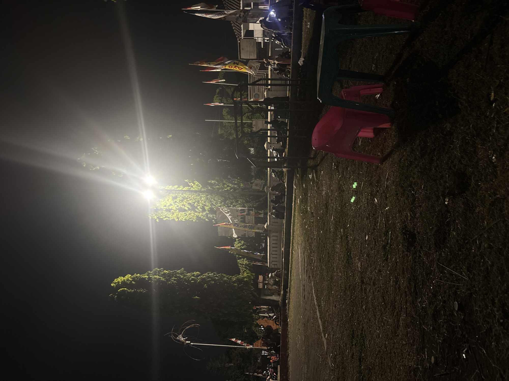 The image captures a nighttime scene in what appears to be a public space in Palimanan, Indonesia, likely a town square or park. A bright overhead light illuminates the area, creating strong highlights and deep shadows, with visible light flares. In the foreground, a pink plastic chair and a green plastic table are present on dry, patchy grass. Further back, a low stone wall runs horizontally, with people sitting and standing behind it. Behind the wall, several trees are visible, some illuminated by the overhead light, and others silhouetted against the dark sky. Various flags, including Indonesian flags and banner-like flags with text and images, are displayed, suggesting a possible event or celebration. Motorcycles are parked in the background, indicating public transport or personal vehicles. Buildings with some signage are also visible in the distance. The overall atmosphere is that of a casual gathering in an outdoor public space at night.