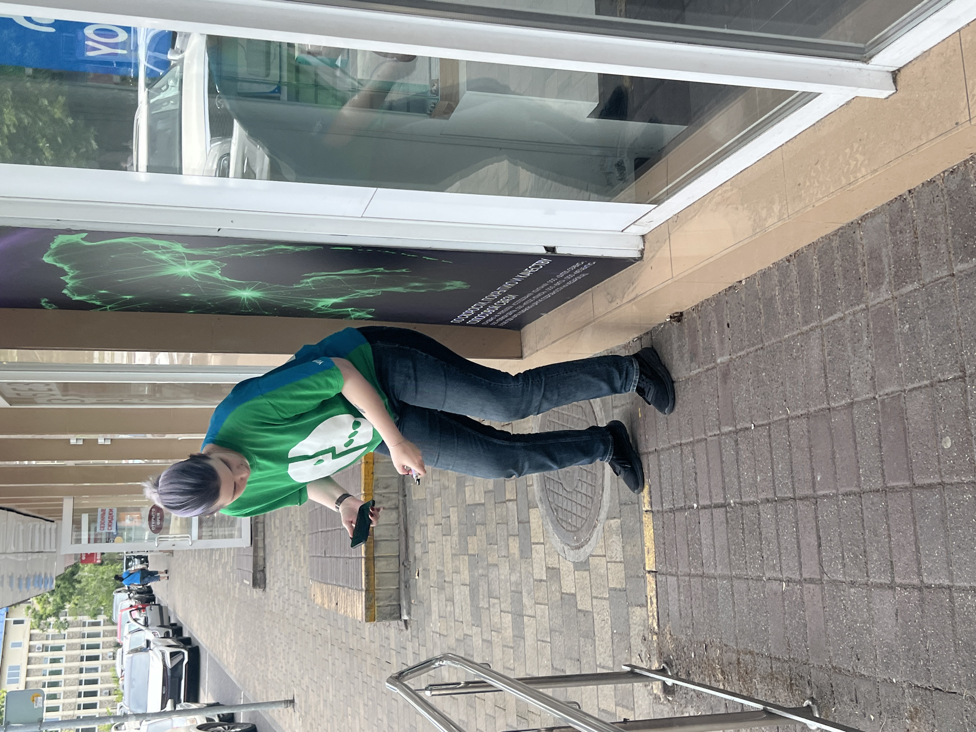 In Sochi, Russia, a person with purple-streaked hair, wearing a green t-shirt with a white logo and dark jeans, stands outside a storefront. The person is looking down at their hands, holding a smartphone in their left hand and a small pink object, possibly a vape pen, in their right. The ground is paved with rectangular bricks, and a metal railing runs along the left side of the image. Behind the person, the storefront has large windows displaying graphics and text in a foreign language. Cars are parked along the street in the background, and buildings are visible further away. The scene appears to be a casual, everyday moment captured on a sidewalk in an urban environment.