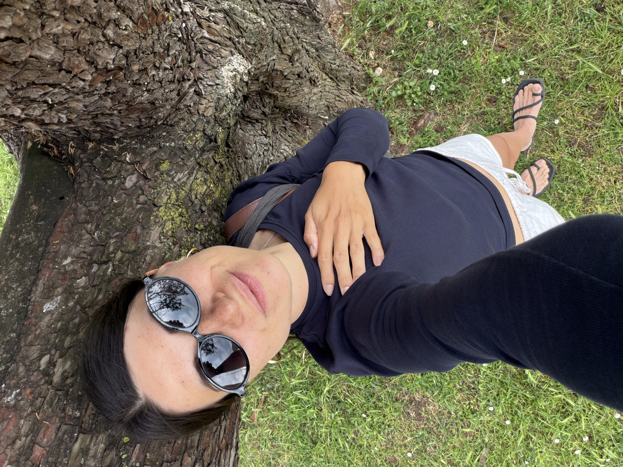 A woman is lying on her side at the base of a tree, likely outdoors on grassy terrain. She has on sunglasses and a long-sleeved dark top. She's wearing light-colored shorts and black sandals. The tree trunk is thick and textured. White flowers are sparsely scattered on the green grass. The specific location in the city of Budva, Montenegro cannot be determined from the visual elements alone.