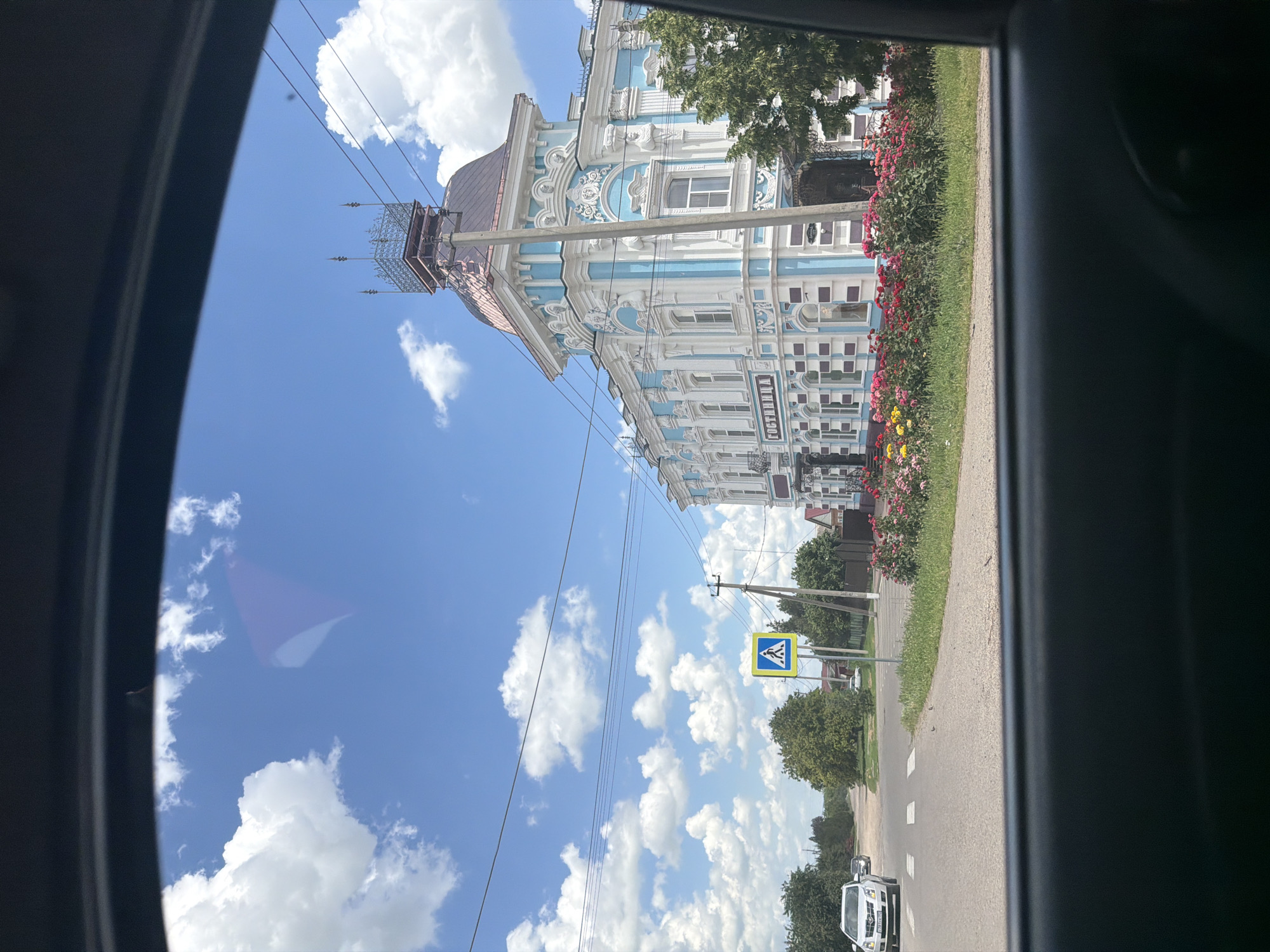 The image, taken from inside a vehicle, captures a view of a street in Leningradskaya, Russia, on a bright, sunny day with scattered white clouds. The camera is tilted, giving a diagonal perspective.
Dominating the scene is a striking, ornate building with light blue and white detailing, featuring intricate architectural elements and numerous windows. A row of vibrant pink and red flowers runs along the front of the building. To the left of the building, a tall pole with what appears to be a metal structure at the top is visible, along with overhead power lines.
On the street in front of the building, a pedestrian crossing sign is prominently displayed on a yellow and blue signpost. The road has white markings indicating lanes and a pedestrian crossing. A few trees line the street, and the overall setting suggests a moderately urban or town environment. A white car is visible on the road, and another white vehicle can be seen further down.
The time of day appears to be midday, given the bright sunlight and clear blue sky. The weather is pleasant and sunny. The word "ГОСТИНИЦА" (Hotel) is visible in large letters on the building, clearly identifying its purpose.
