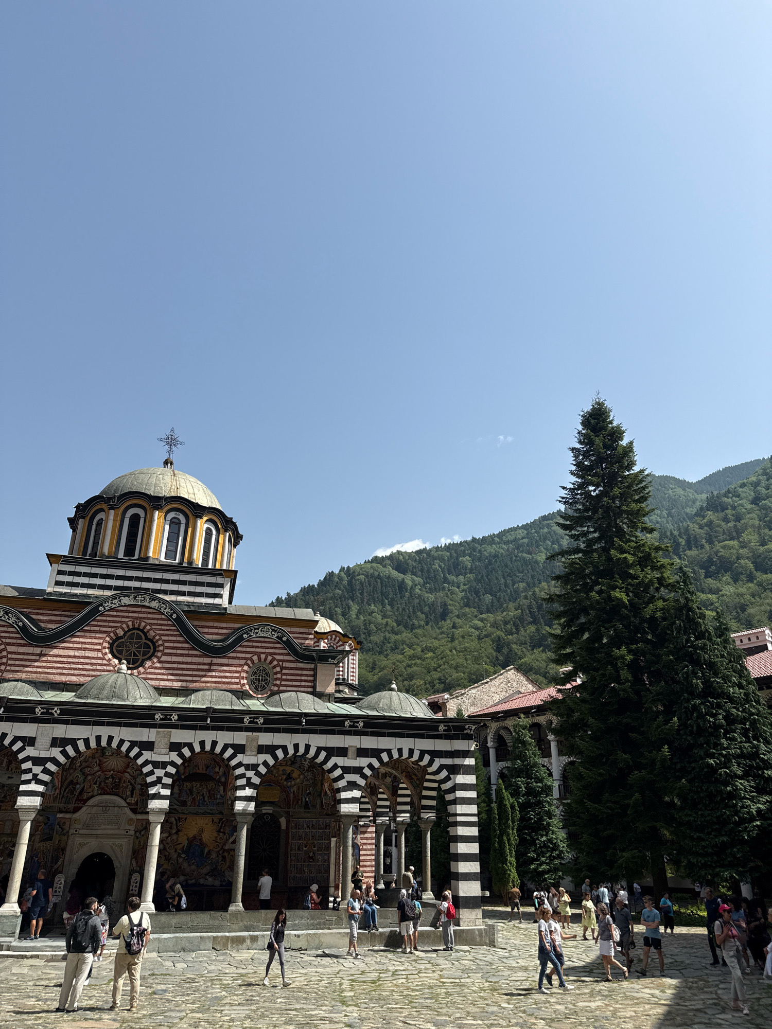 The image shows the Rila Monastery in Rilski manastir, Bulgaria, under a clear blue sky. The monastery's main church, with its distinctive black and white striped arches and a colorful dome topped with a cross, dominates the left side of the frame. Detailed frescoes adorn the walls beneath the arches. In the foreground, a cobblestone courtyard is populated by a diverse group of tourists, dressed in casual summer attire, engaged in various activities such as walking, standing, and observing the architecture. Two tall evergreen trees stand to the right of the church, with a lush, forested mountain range visible in the background. The overall scene depicts a sunny day at a significant historical and religious landmark.
