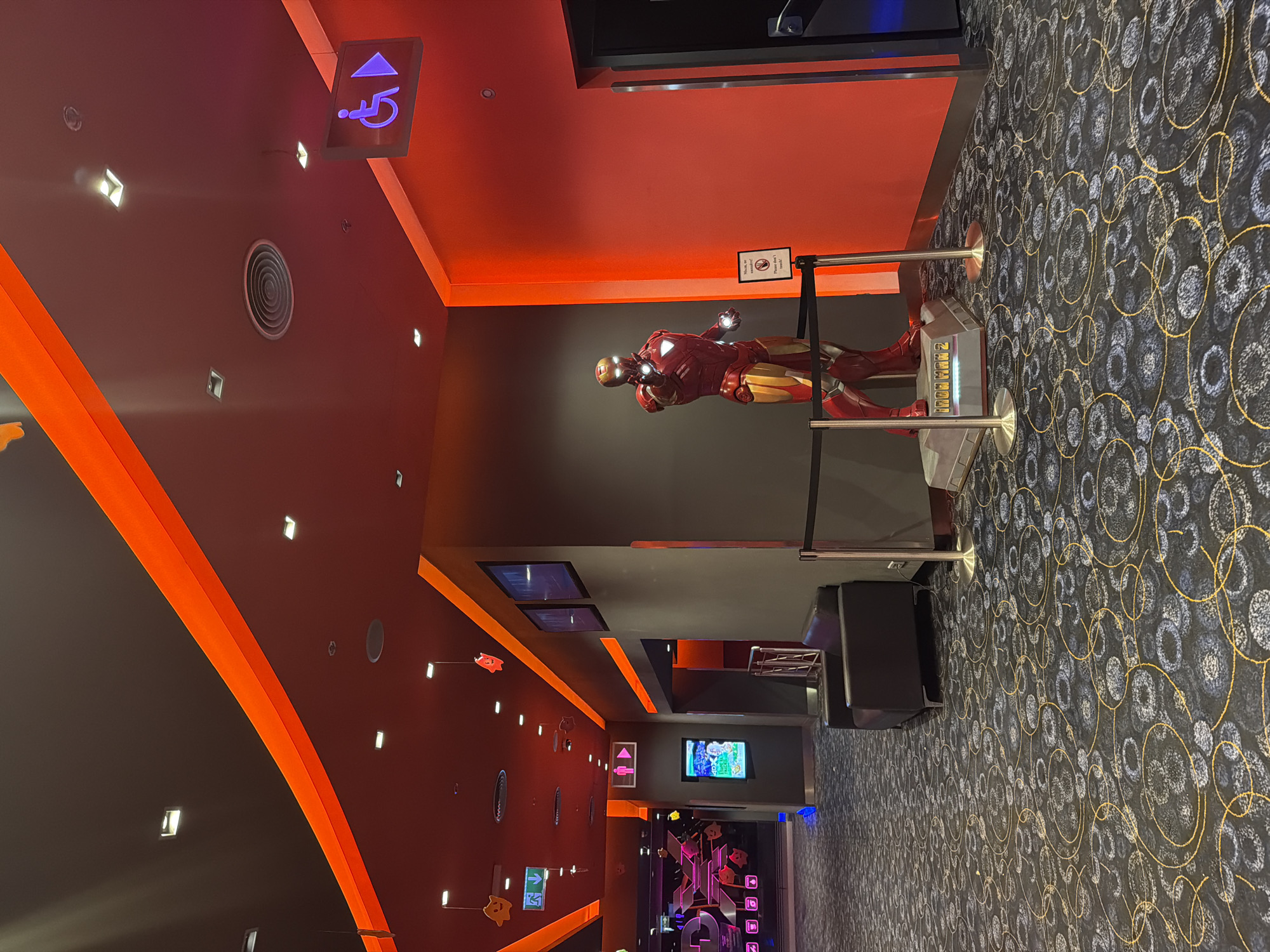 The image captures a scene inside a movie theater, likely in Sofia, Bulgaria, given the information provided. A life-sized statue of Iron Man stands behind a silver barrier. The walls are painted in a grey and orange scheme. The ceiling is dark with recessed lights. Carpet with circular patterns covers the floor. Signage indicates accessible restrooms. The theater appears to be modern, with digital displays in the distance showing movie listings. There are no people visible in the immediate foreground. The focus is on the statue of Iron Man and the ambiance of the theater lobby.