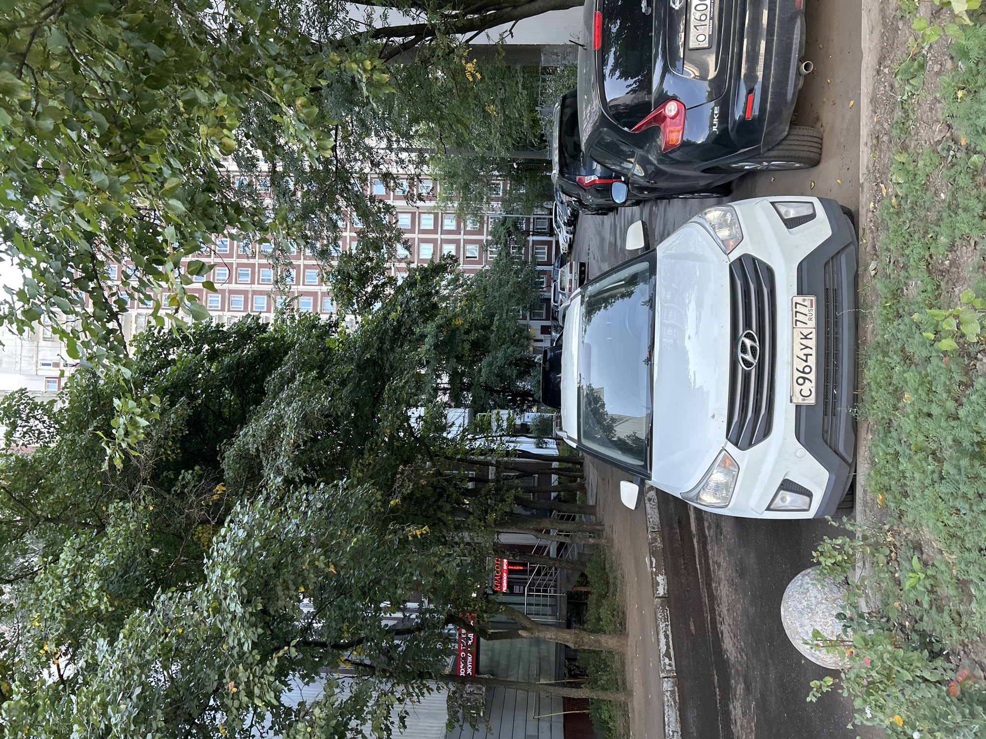 A daytime, eye-level shot shows a street in a residential area with parked cars and trees. In the foreground, a white Hyundai Creta SUV is parked on the right, facing the camera. To its right, a black Nissan Juke is parked, with its rear visible. The license plate of the Hyundai reads "C964YK 777 RUS". A round, speckled concrete bollard is on the left side of the road in front of the Hyundai, with greenery and some plants around it. The road surface is asphalt.
Behind the cars, a row of trees with lush green foliage lines the street, partially obscuring a multi-story residential building. The building is tall, with many windows arranged in a grid pattern and a reddish-brown facade. To the far left, a section of a lighter-colored building with balconies is visible, and a sign with red lettering can be seen. Further down the street, more cars are parked, and the road curves to the right. The sky is overcast, suggesting a cloudy day. The scene appears to be in Moscow, Russia, indicated by the Russian license plate and the architectural style of the buildings.