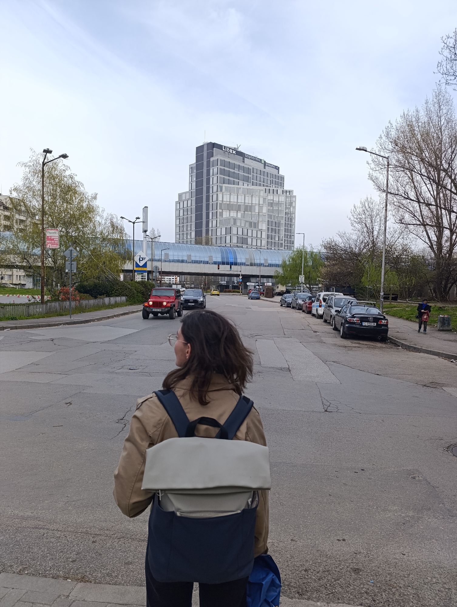 A person with shoulder-length dark hair, wearing glasses, a light brown jacket, and a two-tone backpack (light grey and dark blue), stands at a street intersection in Sofia, Bulgaria. The person is facing away from the camera, looking towards a modern building identified as Litos, with the Sopharma logo visible. Behind the person, a red Jeep is driving on the road. A glass-enclosed pedestrian bridge spans the street, connecting buildings. Cars are parked along the right side of the street. Streetlights and trees are visible along the sides of the road. A pedestrian is walking on the sidewalk on the right. The sky is overcast. The road surface is asphalt with cracks and patches. A sign with red text is visible on a building to the left.