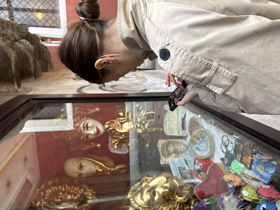 A female-presenting individual, viewed from behind and the right side, stands facing a shop window in what appears to be a narrow thoroughfare in Venice, Italy. The individual has brown hair tied back in a bun and wears a light beige trench coat. Their right hand holds a dark-colored smartphone, displaying an image with light-colored patterns (possibly flowers), while their left hand's fingers are also positioned near the screen, suggesting interaction with the device.
The shop window displays various items, including multiple ornate Venetian masks. Prominently visible are several gold-toned masks: a lion's head mask on the lower left, and a classical Venetian mask and a jester-style mask with a white face and red and gold accents on a red background in the upper section. The display also includes smaller decorative objects, such as sculpted faces, round plaques, and a collection of colorful, patterned, soft pouches or coin purses in the lower left. Reflections are visible on the window glass, including parts of the street scene and possibly the person's own reflection or items from inside the shop.
To the right of the person, a textured building wall is visible, made of plaster or stone, with graffiti-like markings. Above the wall, an older window with a grid pattern and a soft yellow light source inside is present, indicative of the historic architecture common in Venice. The overall setting, characterized by the traditional Venetian masks and architectural style, firmly places the scene in a commercial area within Venice.