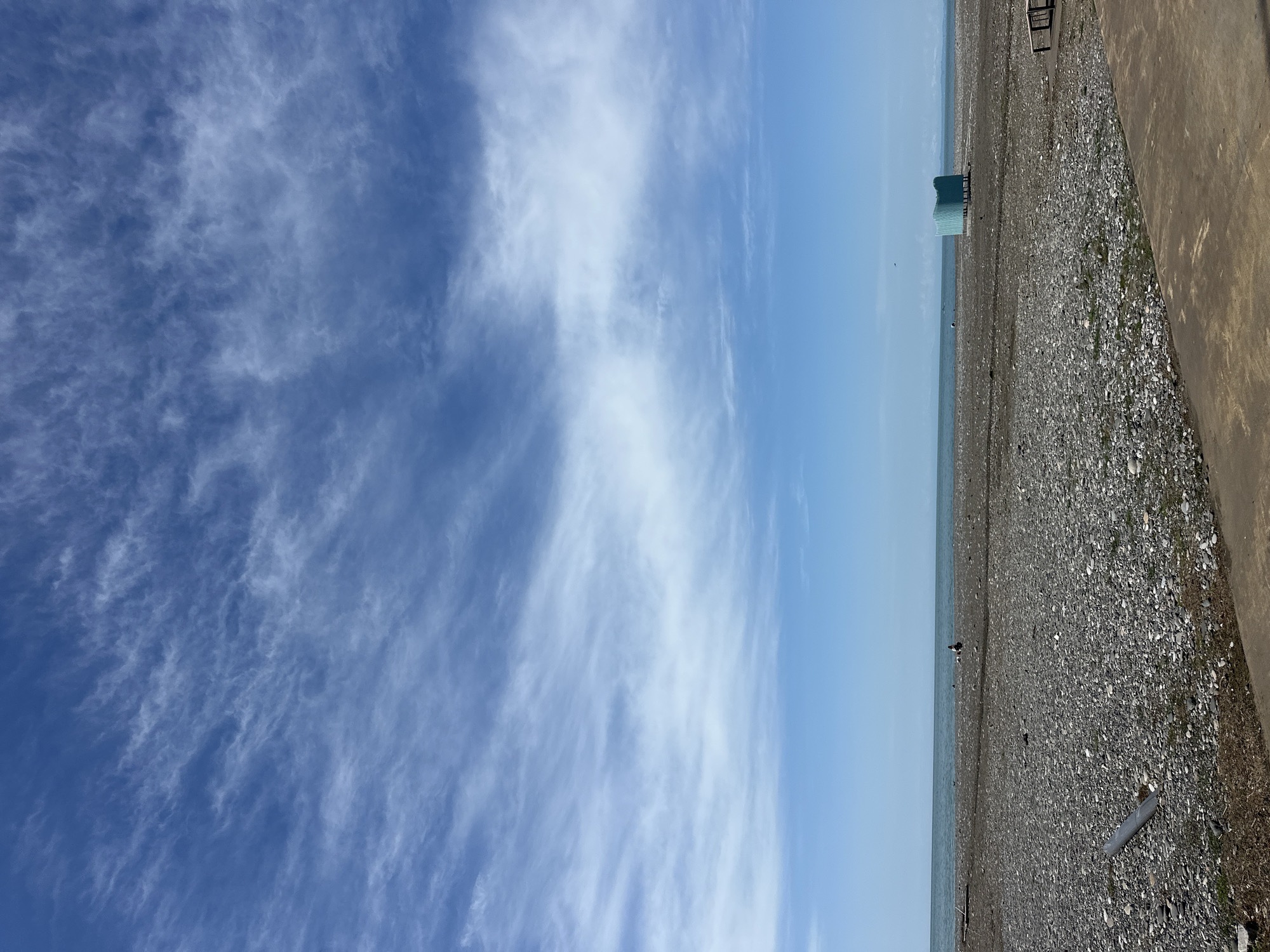 The scene is a shoreline in Batumi, Georgia. The sky is blue with wispy clouds. To the right of the frame, there is a shingle beach composed of small pebbles. A concrete wall runs along the edge of the beach. Two green shelters are located near the sea, on the left of the beach.
A small group of people can be seen towards the sea.