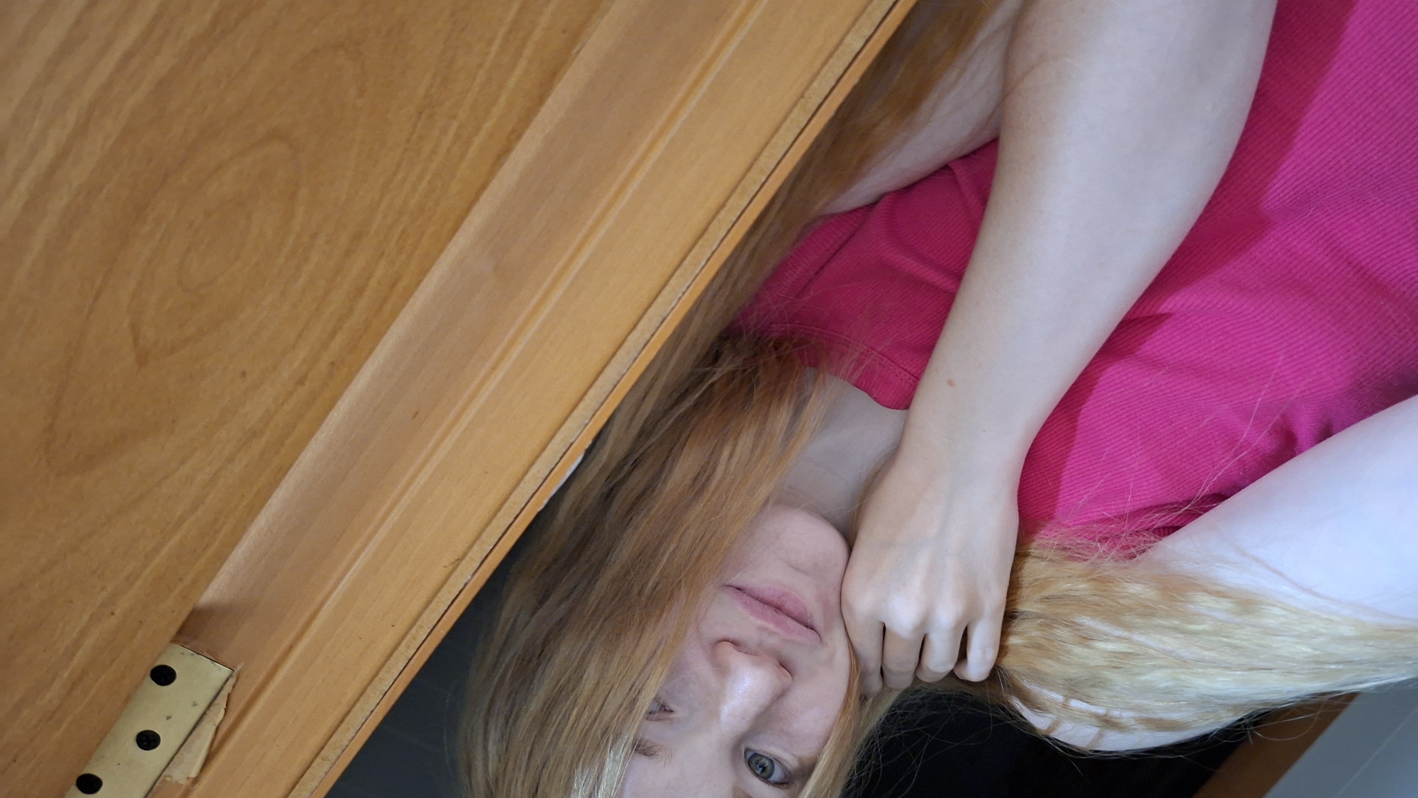 An indoor, close-up image captures a person, likely female, with long, light blonde hair looking up towards the viewer. She is wearing a bright pink t-shirt. Her left hand rests near her chin, gently holding strands of her hair. Her expression is neutral to slightly curious, with her blue-grey eyes open.
A warm-toned wooden surface, possibly a piece of furniture like a desk lid or a cabinet door, diagonally crosses the upper-left portion of the frame. This wooden surface displays visible grain patterns, including an oval-shaped knot. A brass-colored metal hinge with three dark screws is partially visible in the lower-left corner, affixed to the wooden surface. The perspective suggests the person is lying down and looking up, or the camera is positioned above her in a tight space. The overall lighting is soft, indicating an indoor setting. No text is visible in the image. The provided location context for this image is Большой Букор, Russia.